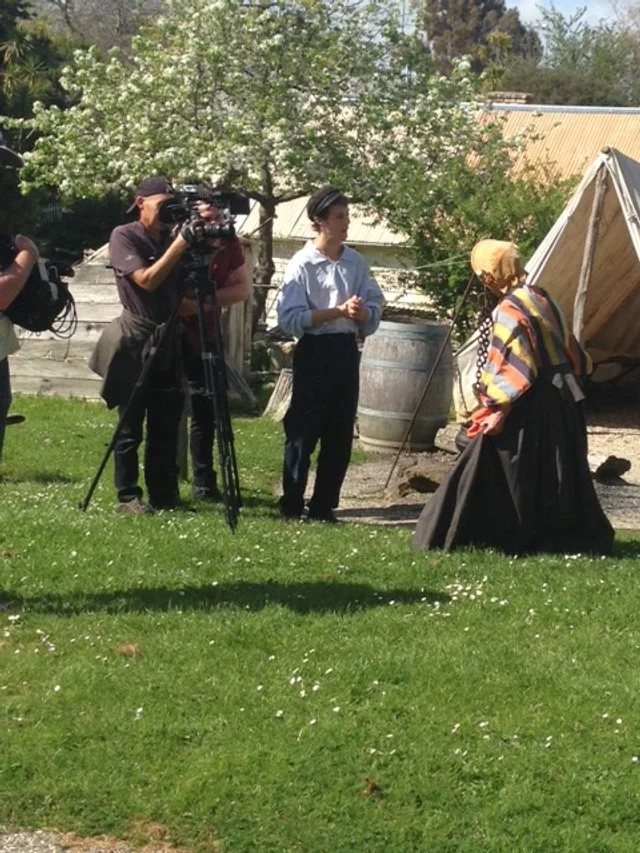A woman dressed in historical clothing being filmed by a cameraman and a crew, outdoors in a garden area with grass, trees, and a barn.