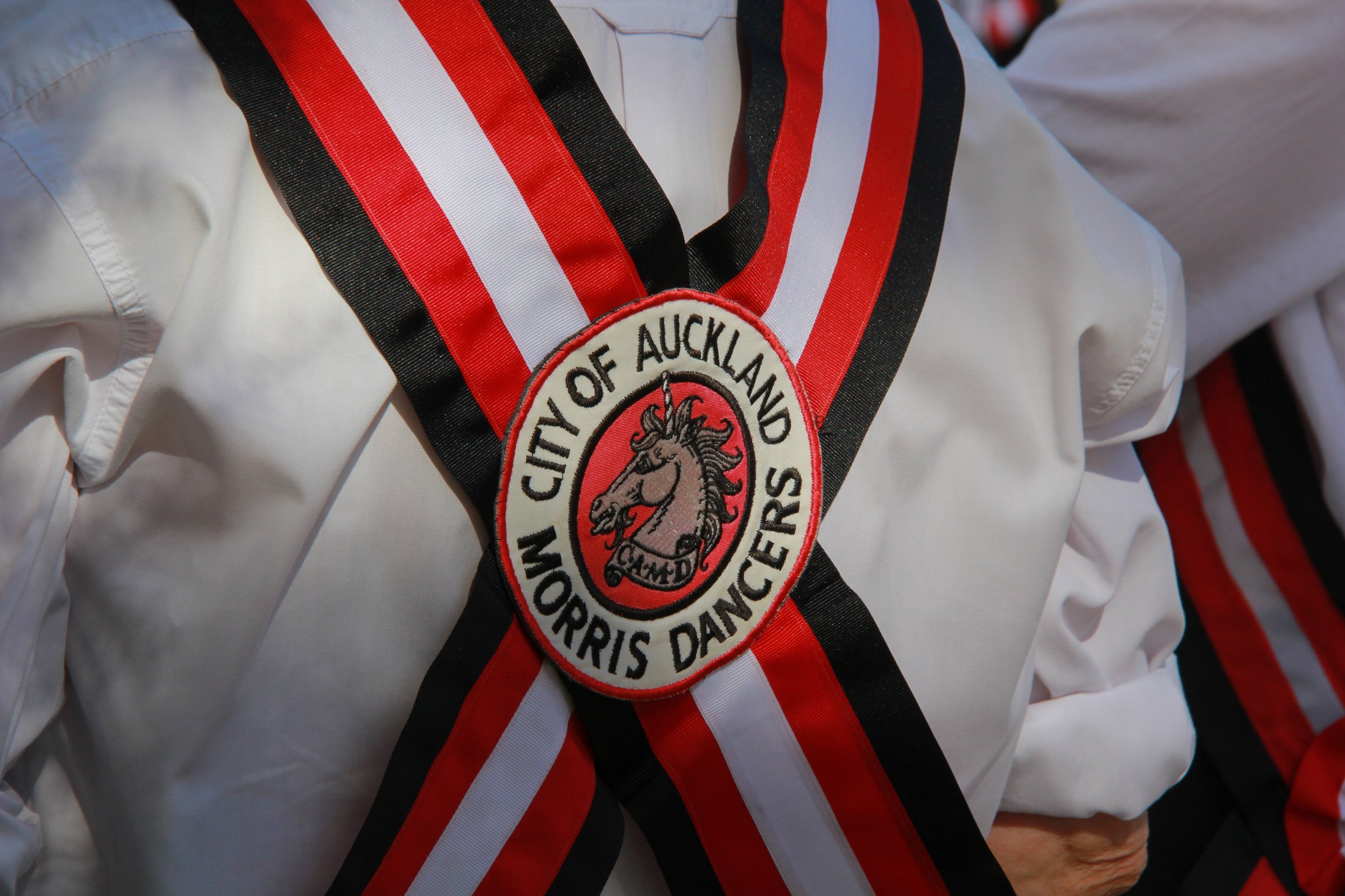 A white chef's jacket with a circular patch that reads 'WOM City of Auckland Morris Dancers.' The patch has a depiction of a unicorn's head at the center. The jacket is decorated with red, black, white, and silver ribbons.