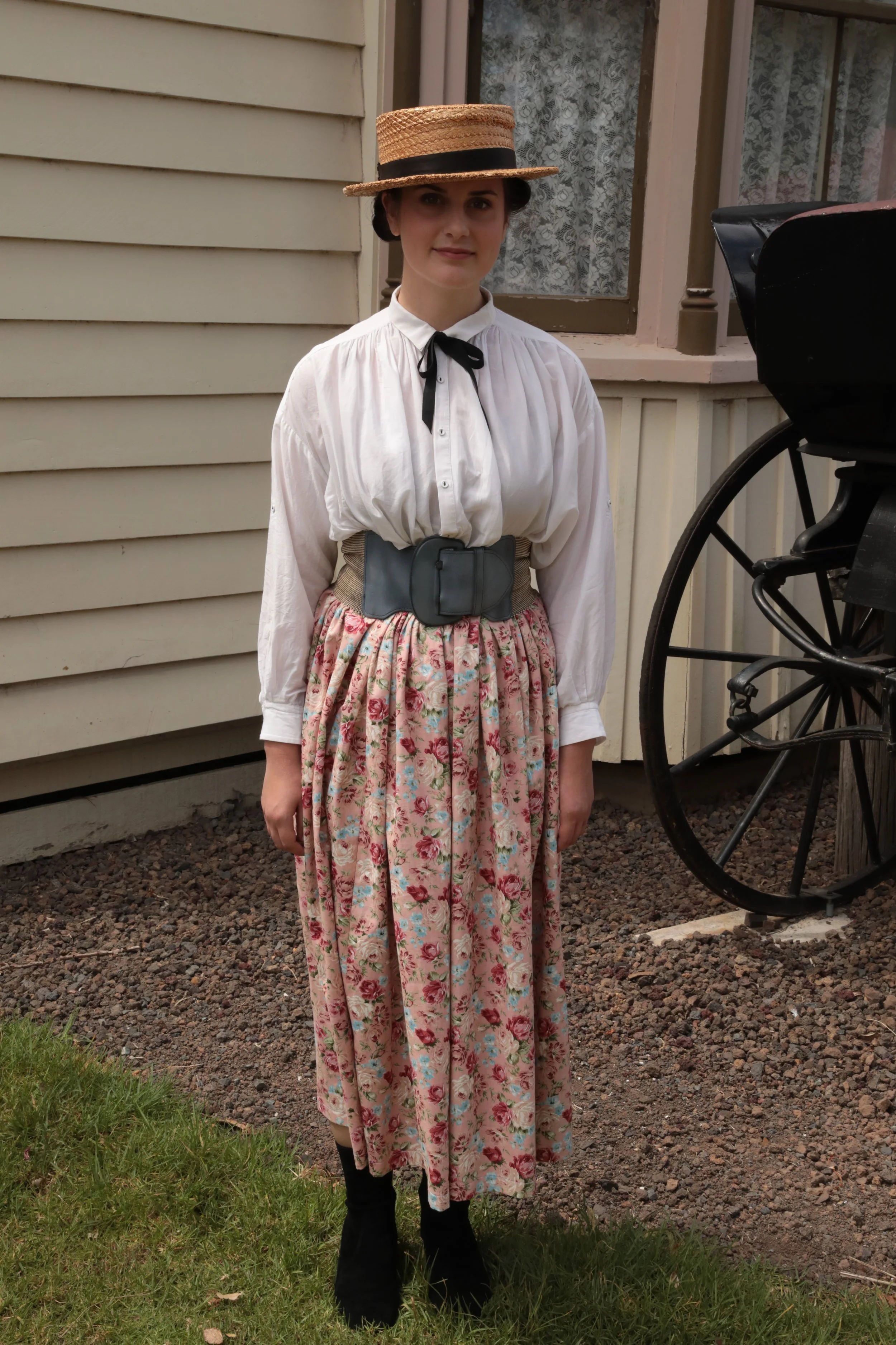 Woman standing outdoors dressed in historical clothing, wearing a wide-brimmed straw hat, a white blouse with a black ribbon, a wide belt, a floral patterned skirt, and black boots.