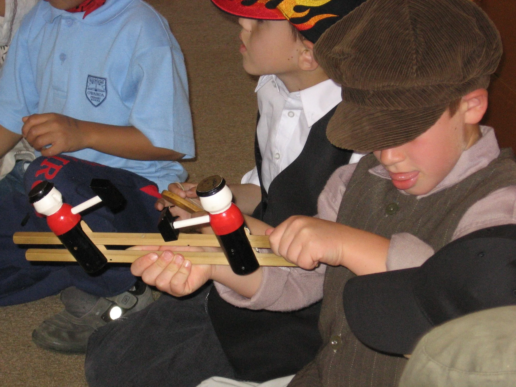 Children sitting on the floor, some are holding small wooden objects and a game resembling a tic-tac-toe setup with red and black pieces. The children are wearing various clothing, including school uniforms and casual outfits, and are engaged in an a