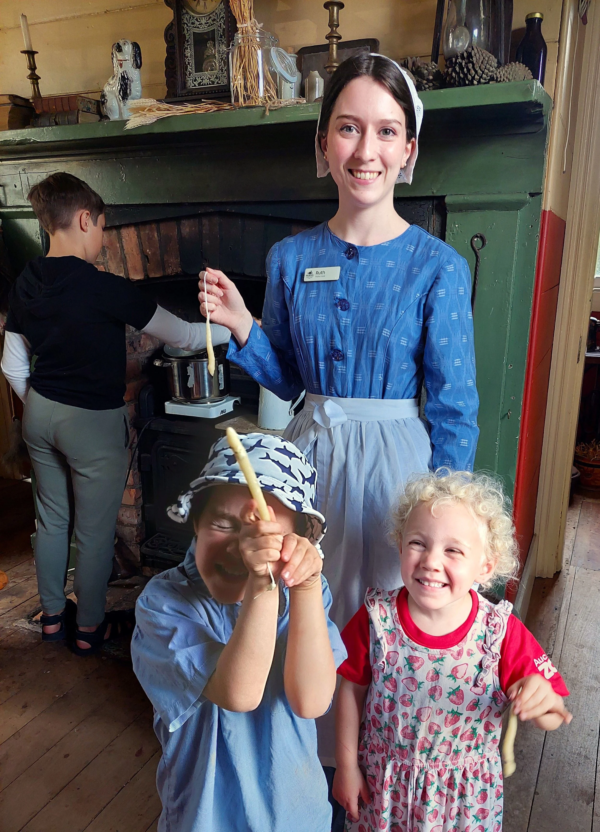 A smiling young woman dressed as a historical figure, possibly from the 1800s, stands in front of a green vintage fireplace. She is wearing a blue dress with a white apron and a white bonnet. Around her are children, two of whom are holding long, lig