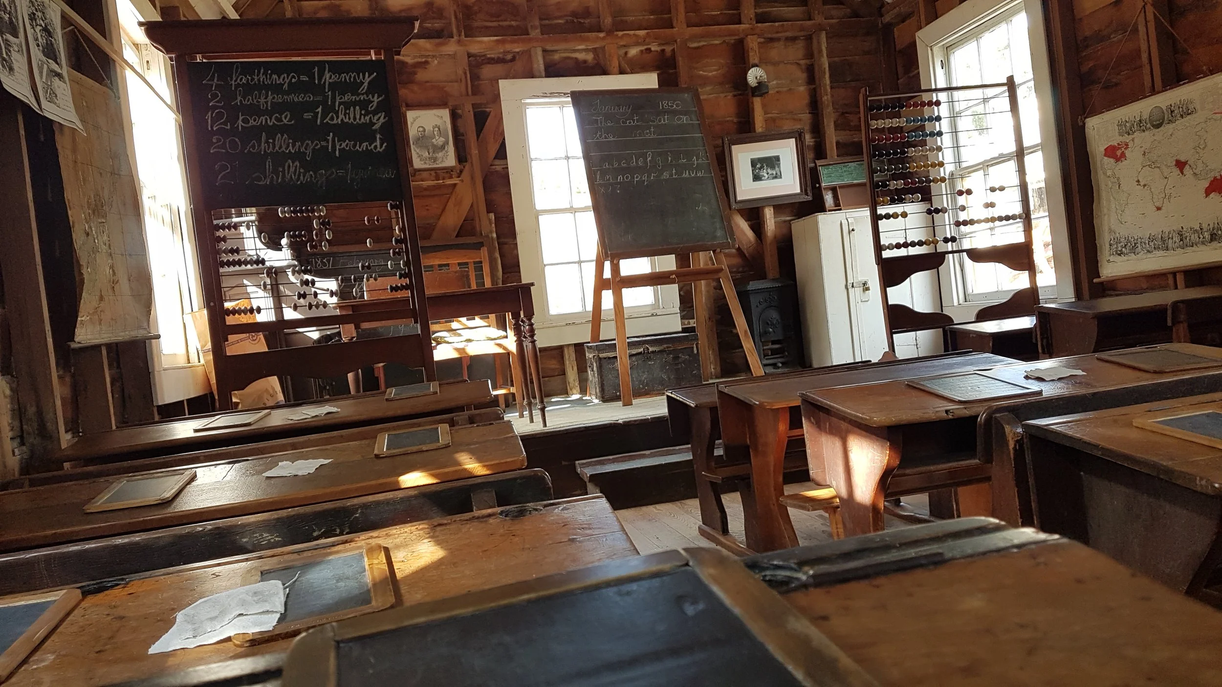 An old-fashioned classroom with wooden desks and chairs, large windows letting in sunlight, and chalkboards with writings and drawings on the walls.
