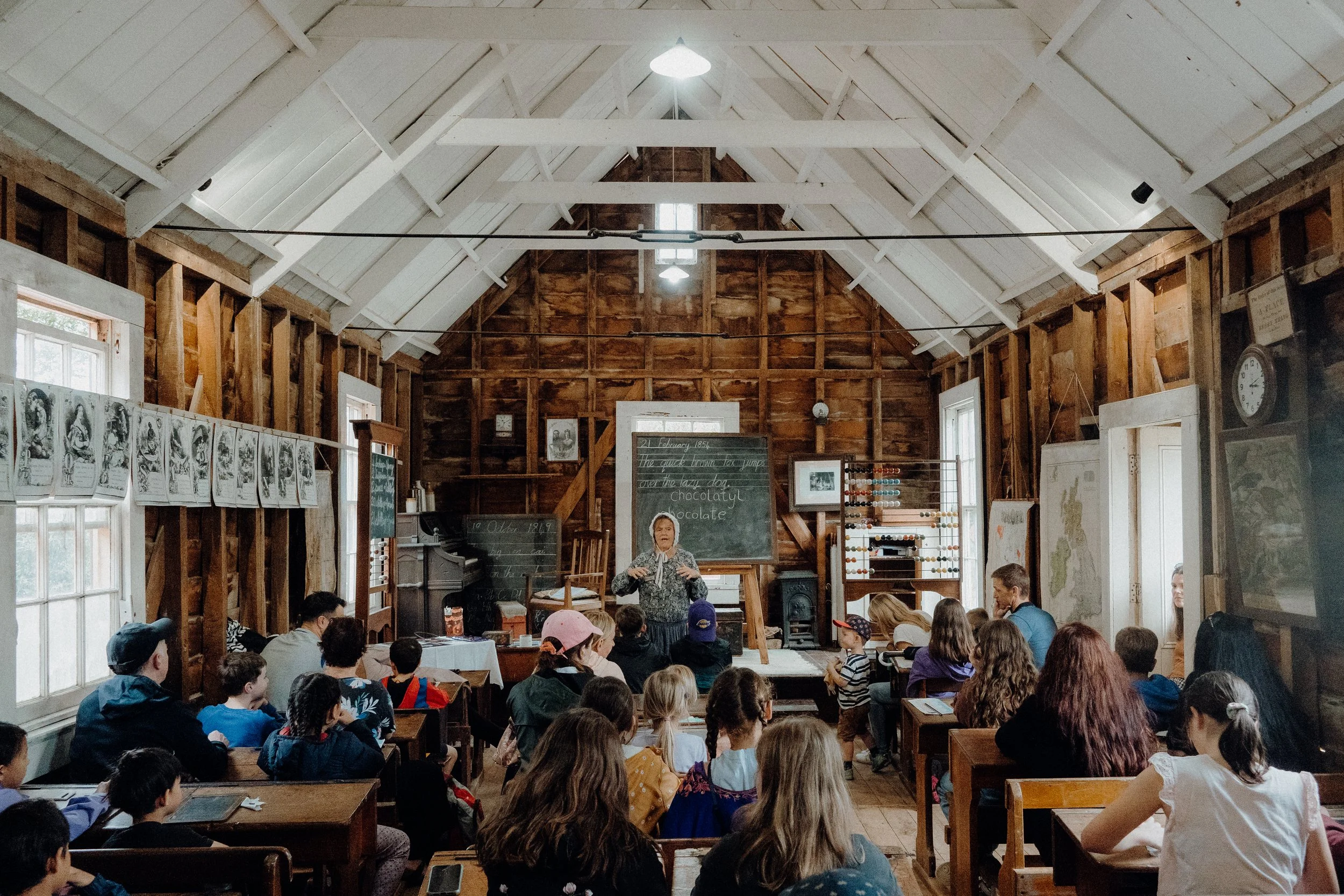Children sitting at desks in a vintage-style classroom with exposed wood walls and a high ceiling, listening to a woman in front of a chalkboard with writing and a map hanging.