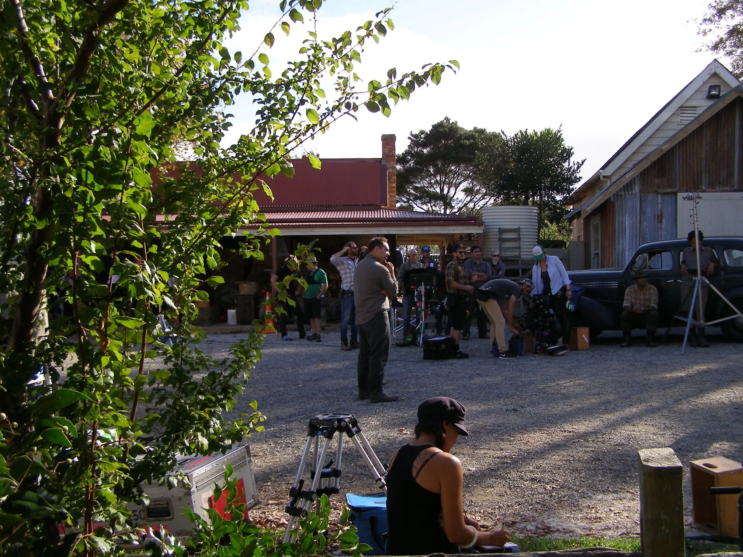 Film crew preparing equipment outdoors with people gathered around, vintage truck, rustic buildings, and trees in the background.