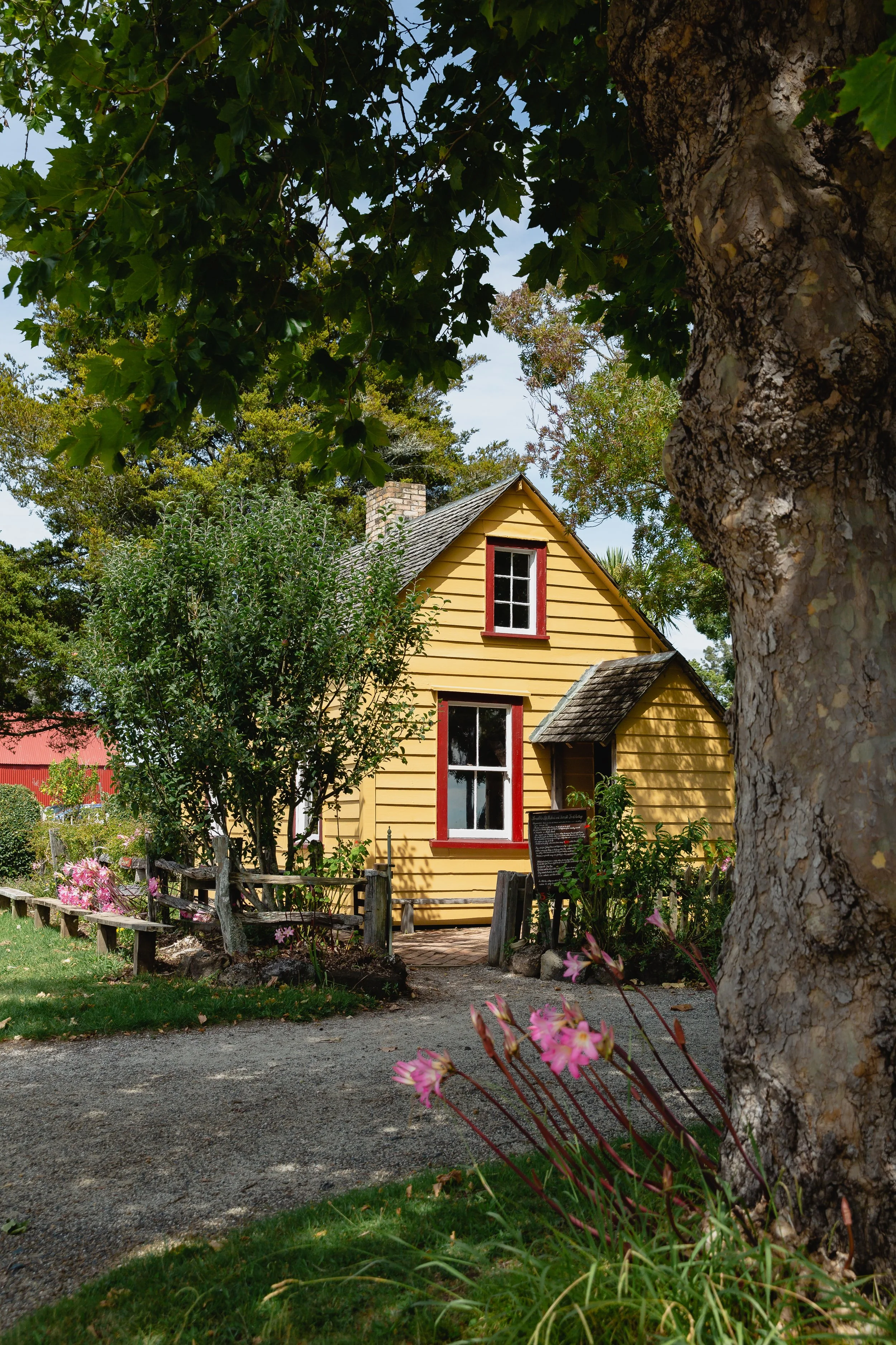 A small yellow wooden house with red window frames, surrounded by trees and flowers, viewed from a fairy-tale-like garden pathway.