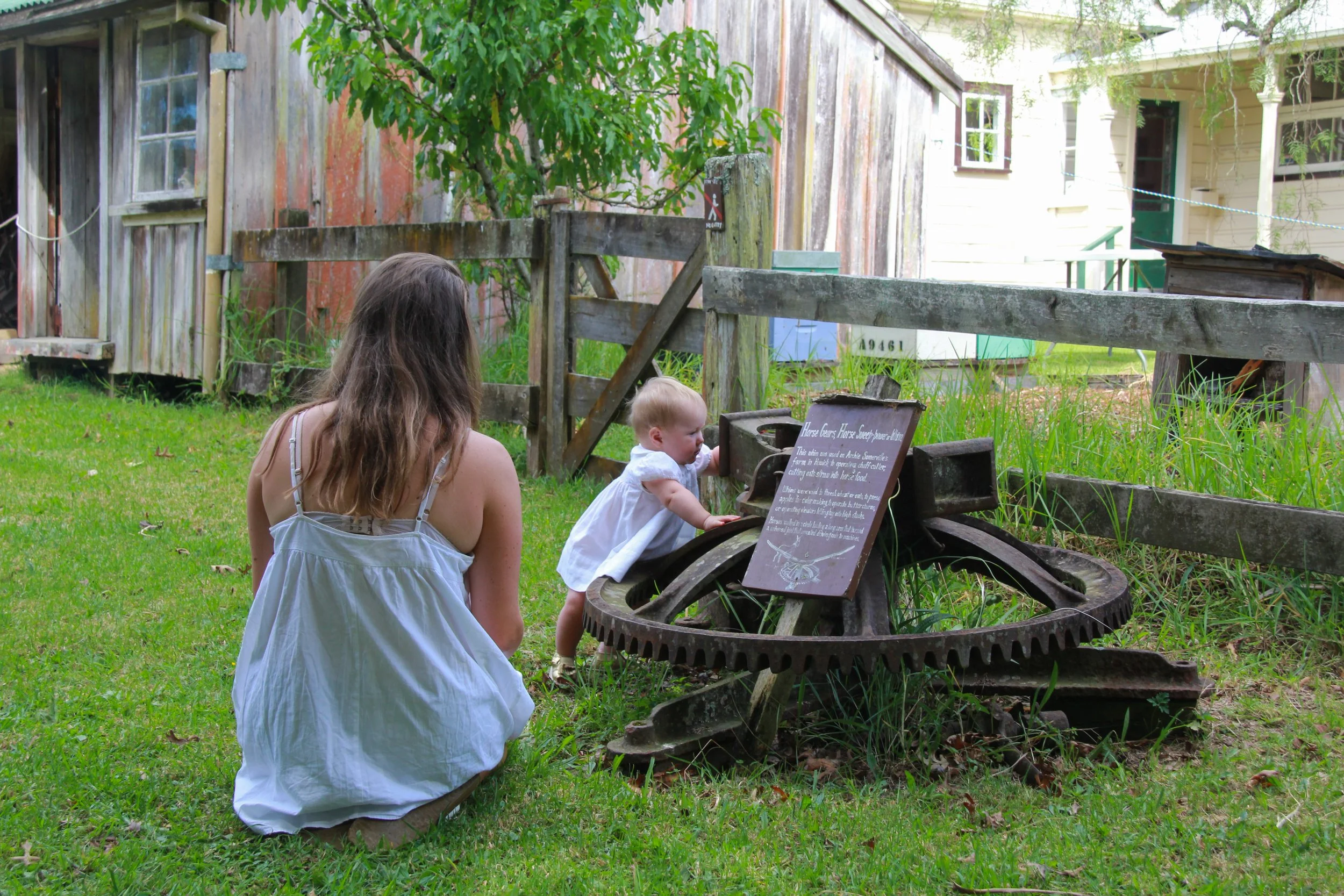 A woman and a young girl examine a historical artifact outside, with aged wooden buildings and a green yard in the background.