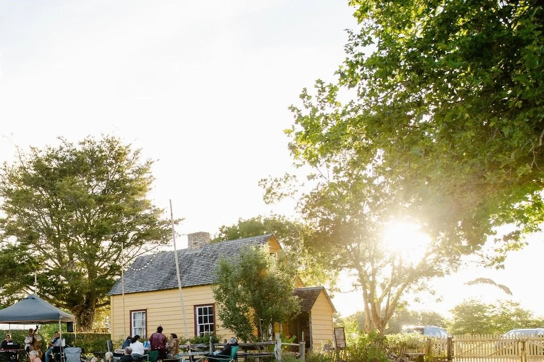 Yellow cottage surrounded by trees with people gathered outside, sun shining through foliage, and a picket fence in the background.