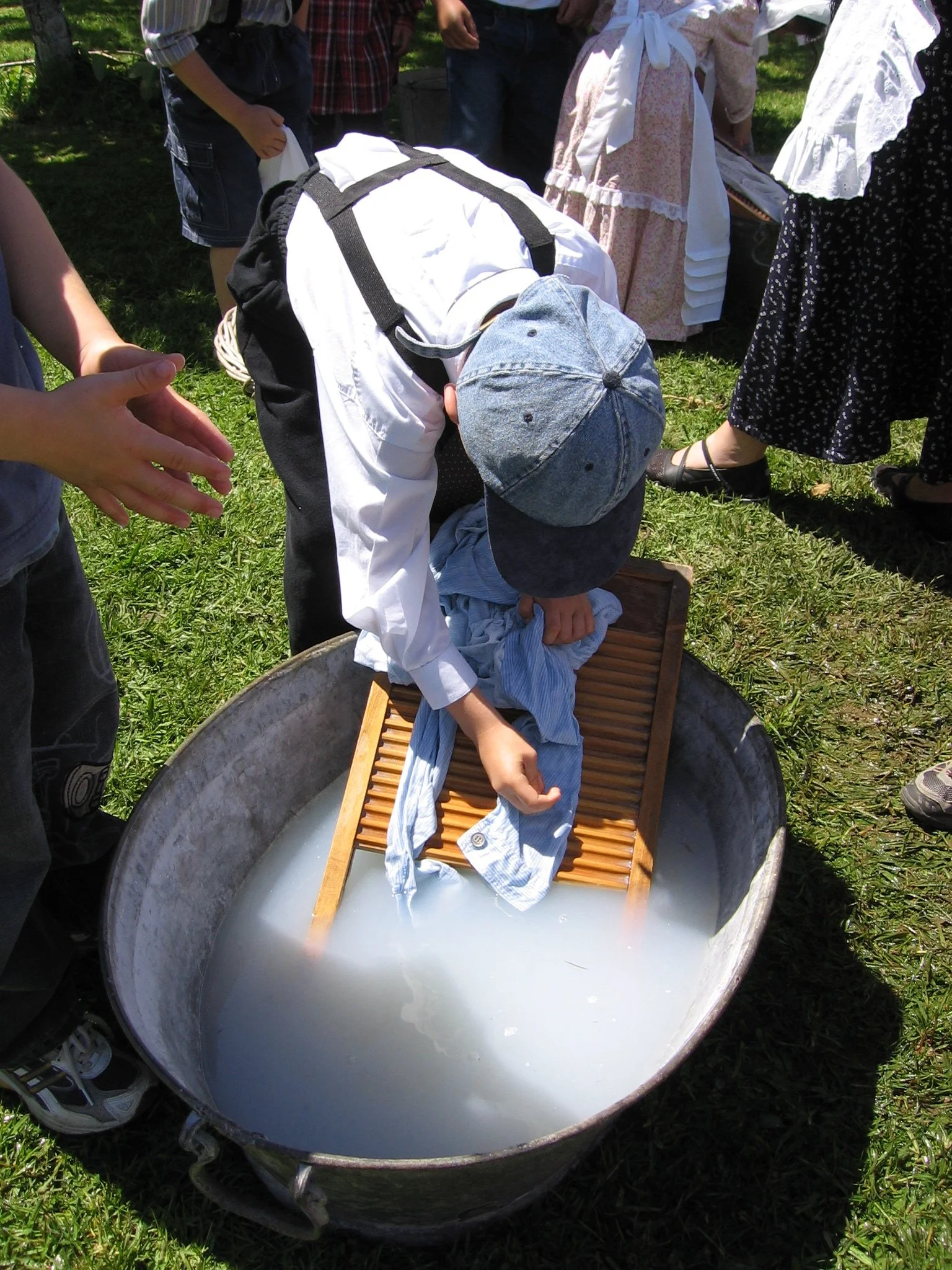People gathered around a large metal tub filled with water. A child in a denim cap and white shirt is leaning over a washboard placed inside the tub. Several adults stand nearby, some dressed in traditional or vintage-style clothing, outdoors on a grassy area on a sunny day.