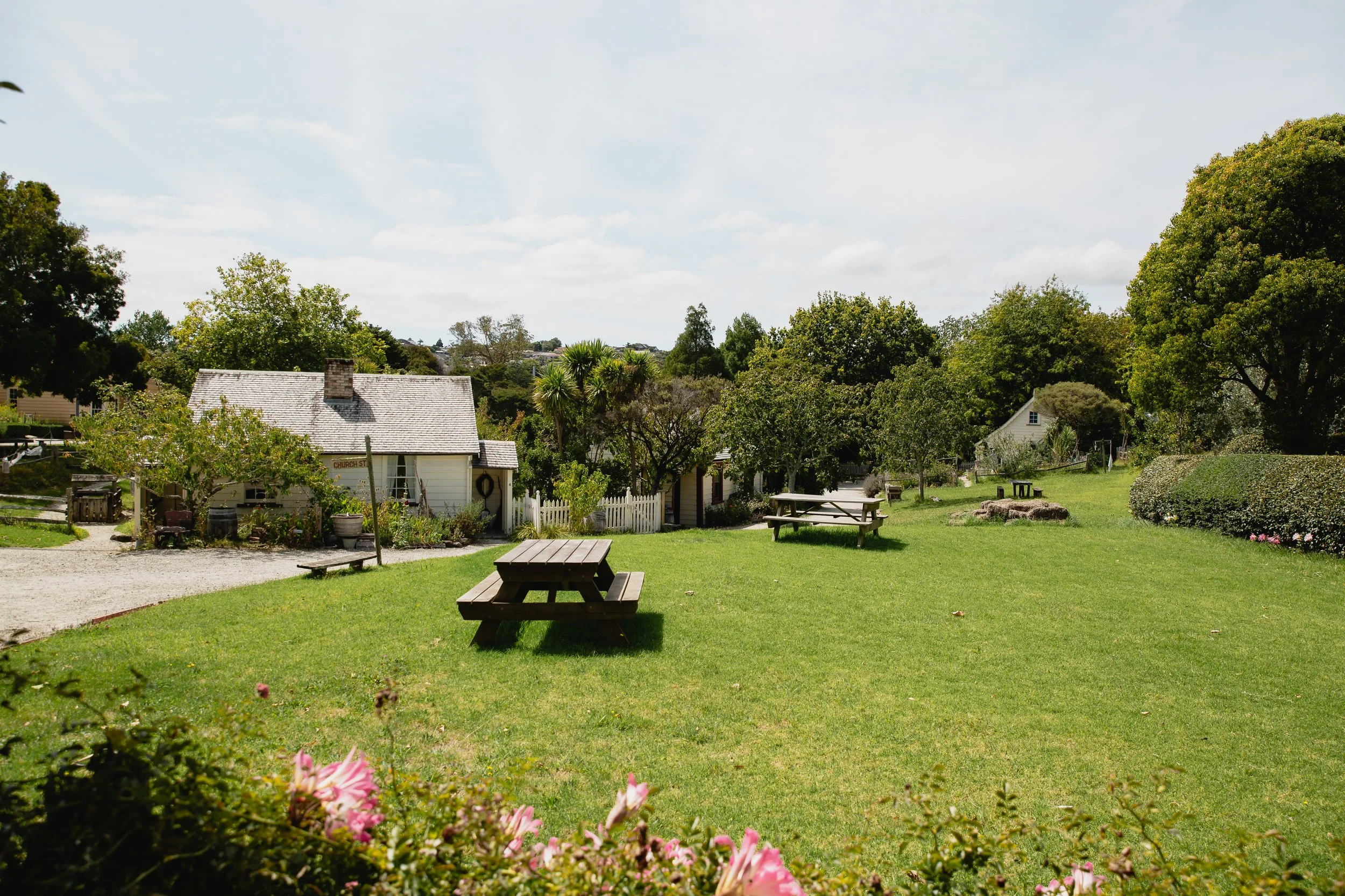 A green grassy yard with several picnic tables, surrounded by trees and small houses under a partly cloudy sky.
