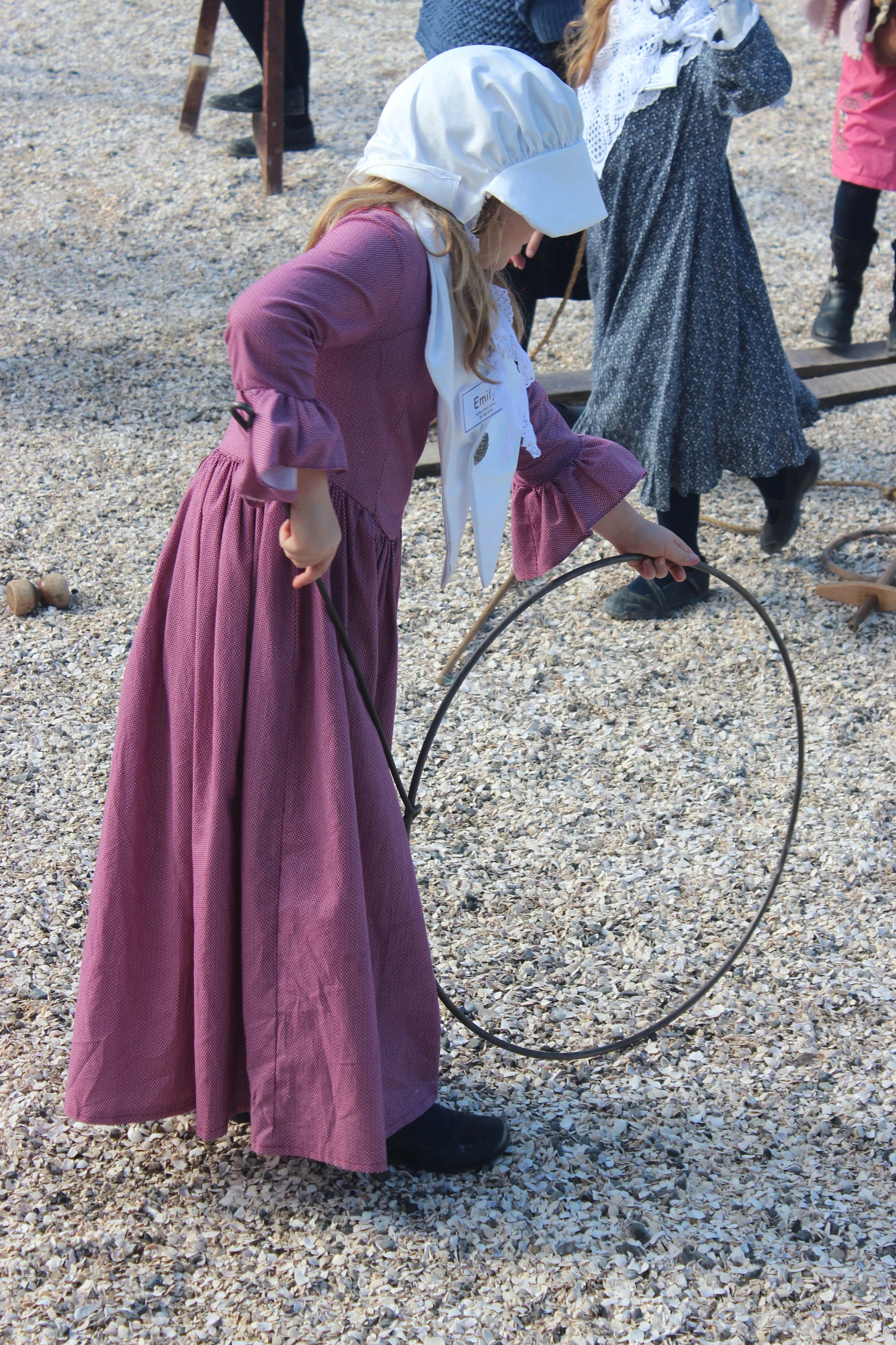 A young girl dressed in historical clothing, with a name tag that says 'Emily,' is holding a hoop and standing on a gravel surface at an outdoor historical event.