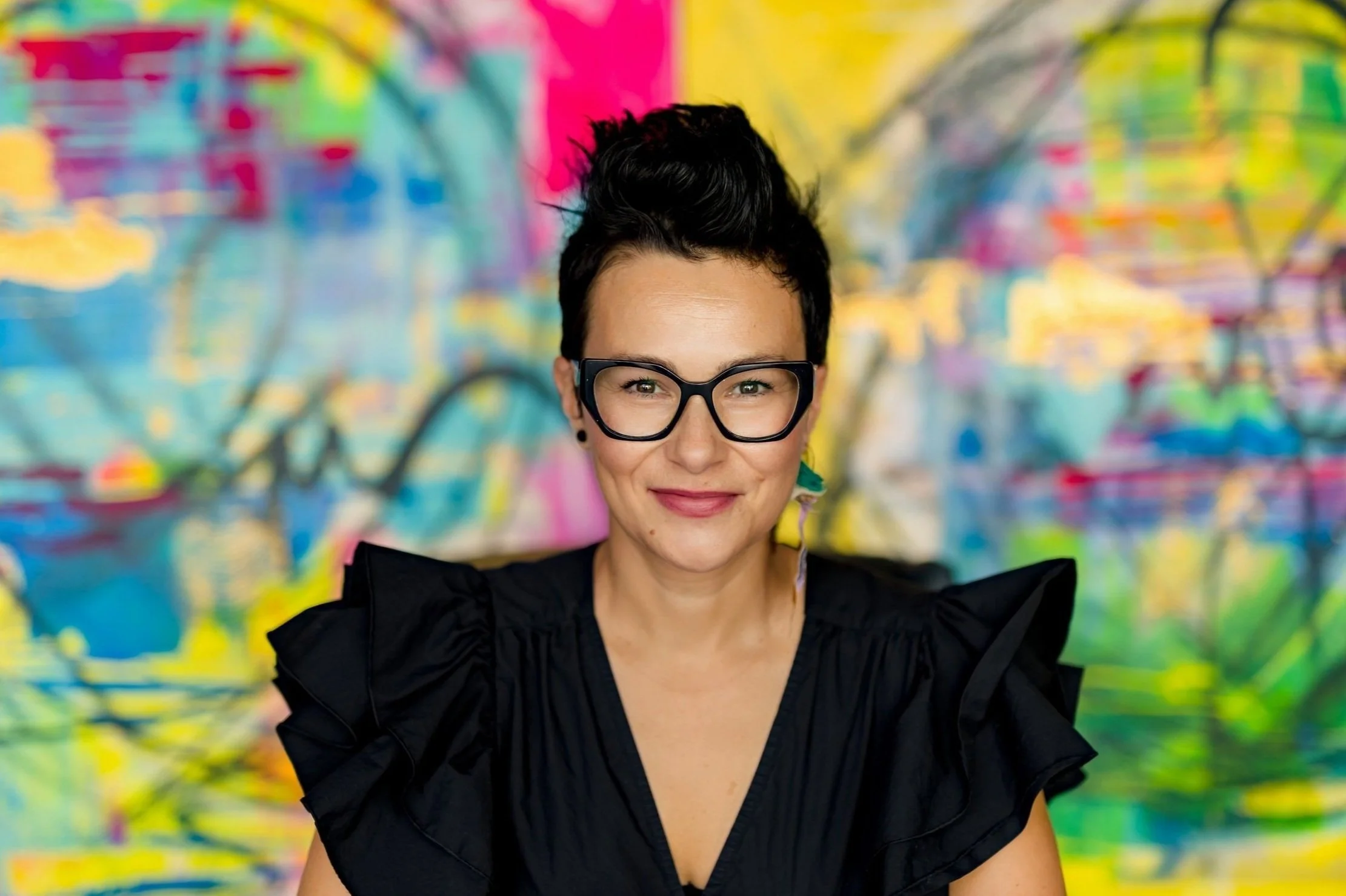 Portrait of a woman with short dark hair, black glasses, and wearing a black ruffled top, standing in front of a colorful abstract mural.