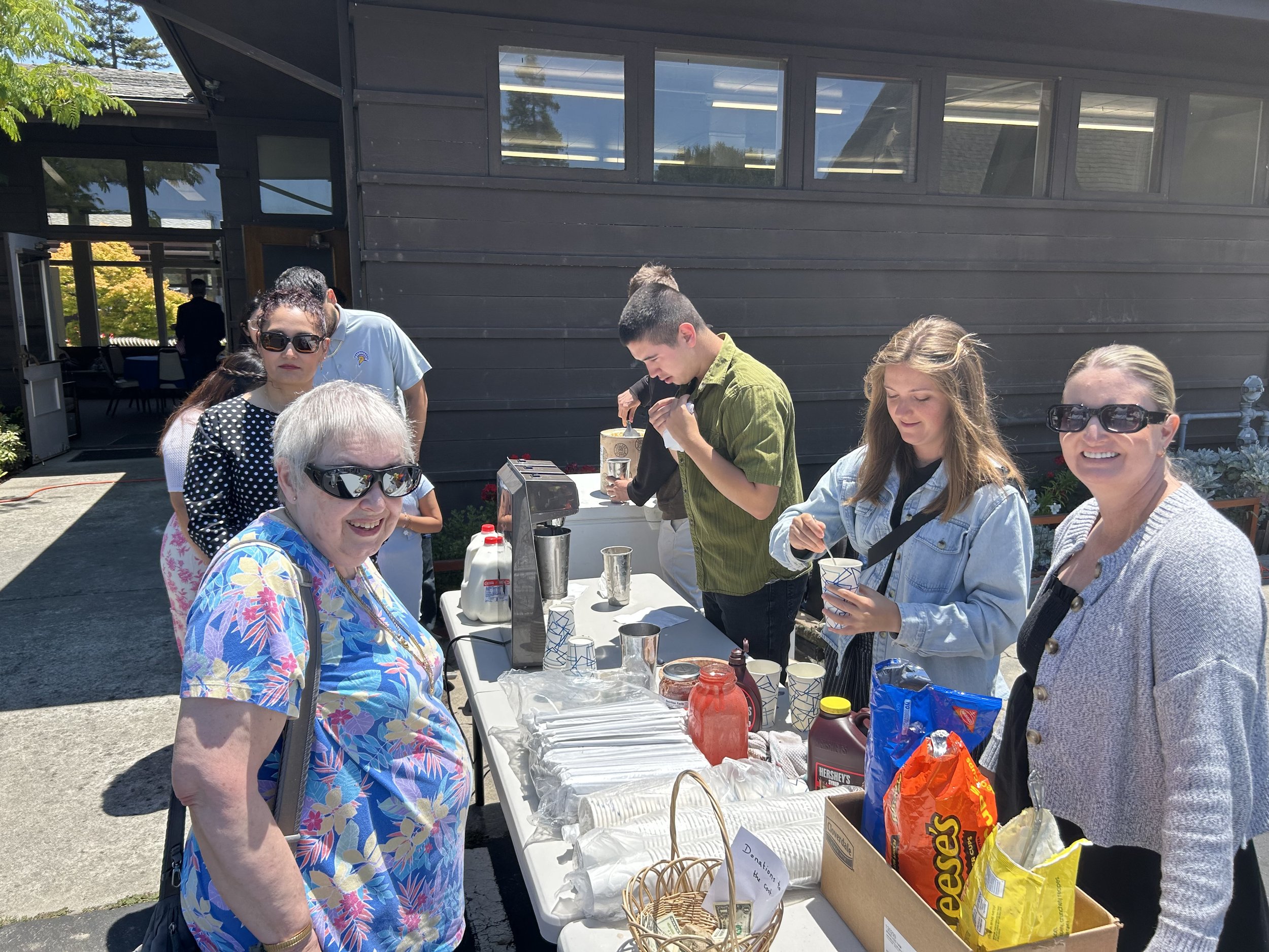 People gathered around a table outside, serving ice cream and snacks on a sunny day.