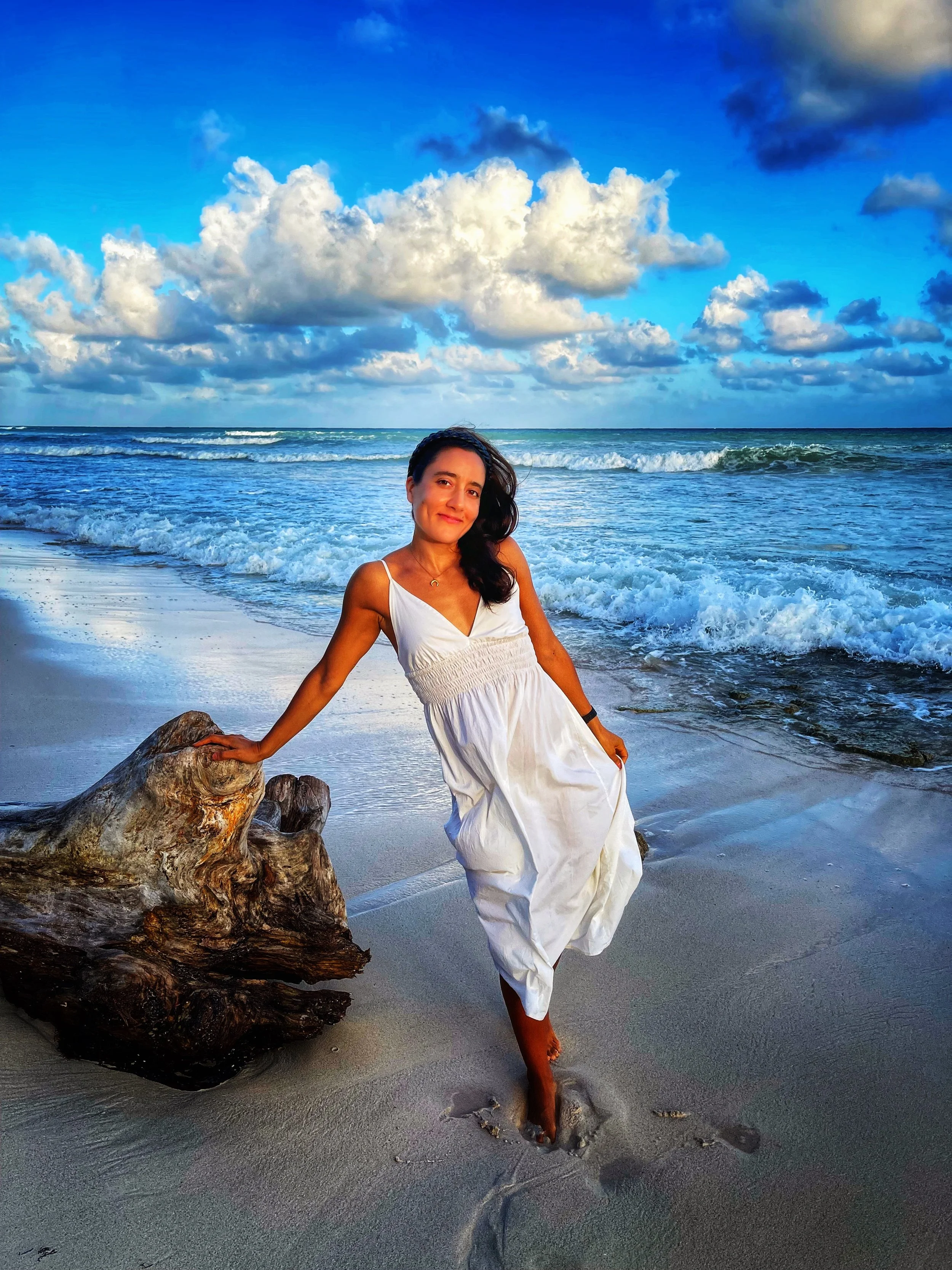 Mujer con vestido blanco en la playa, tocando una pieza de madera, con el mar y nubes en el cielo de fondo.