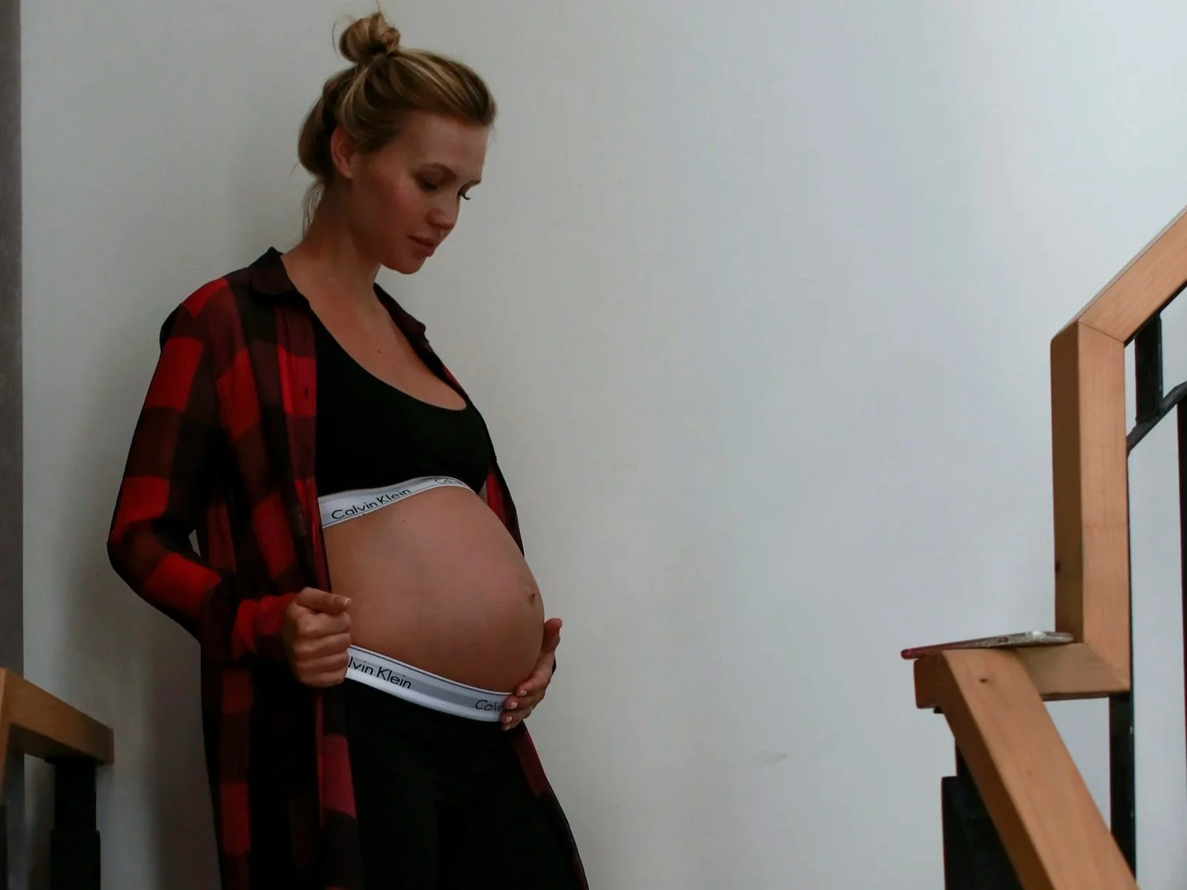 A pregnant woman paused halfway up a wooden staircase, looking down lovingly at her belly, symbolizing the transitional journey of matrescence and the ascent into her own power as the authority of her birth.