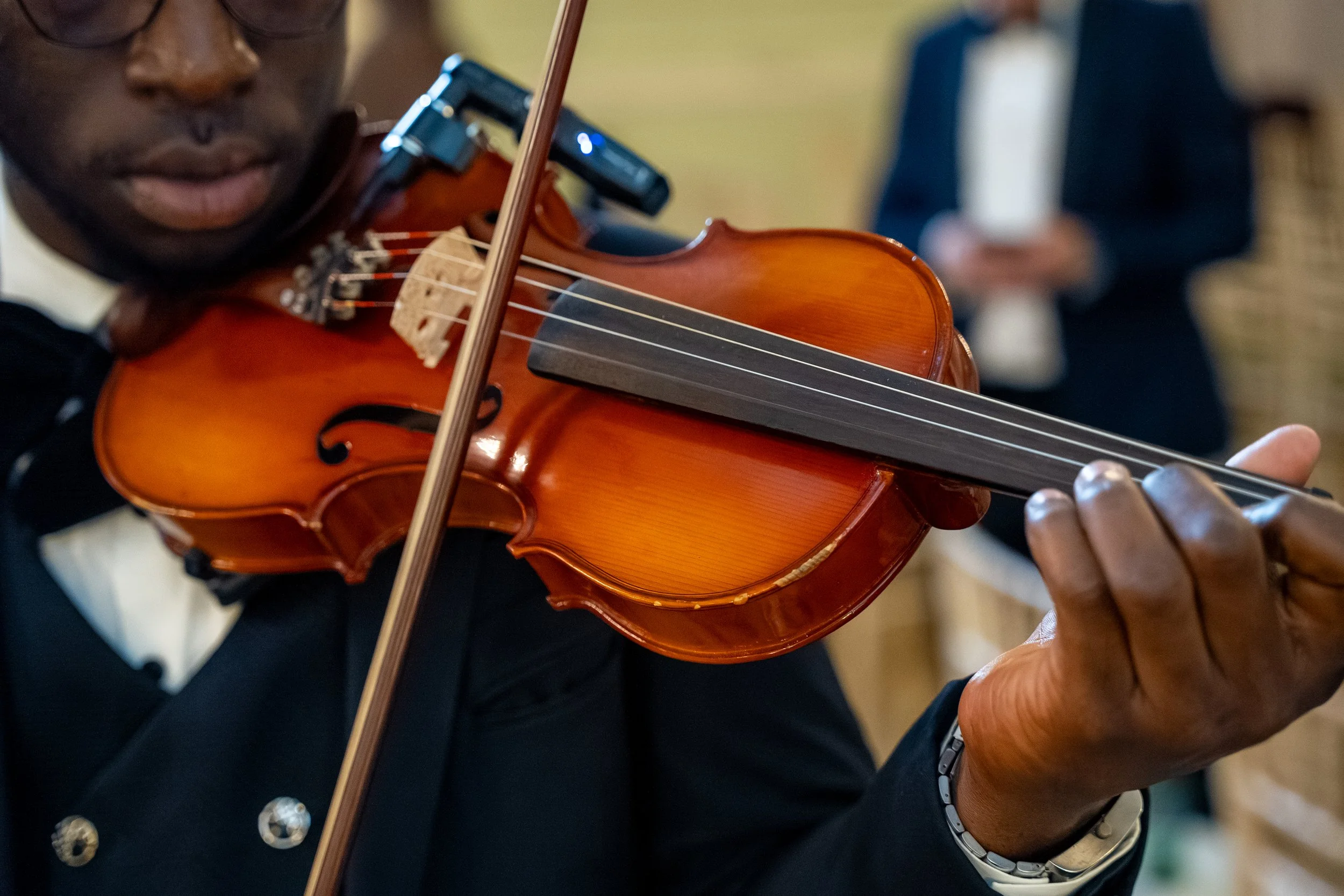 Close-up of a person playing a violin, focusing on the instrument and hands, with a blurred person in the background.