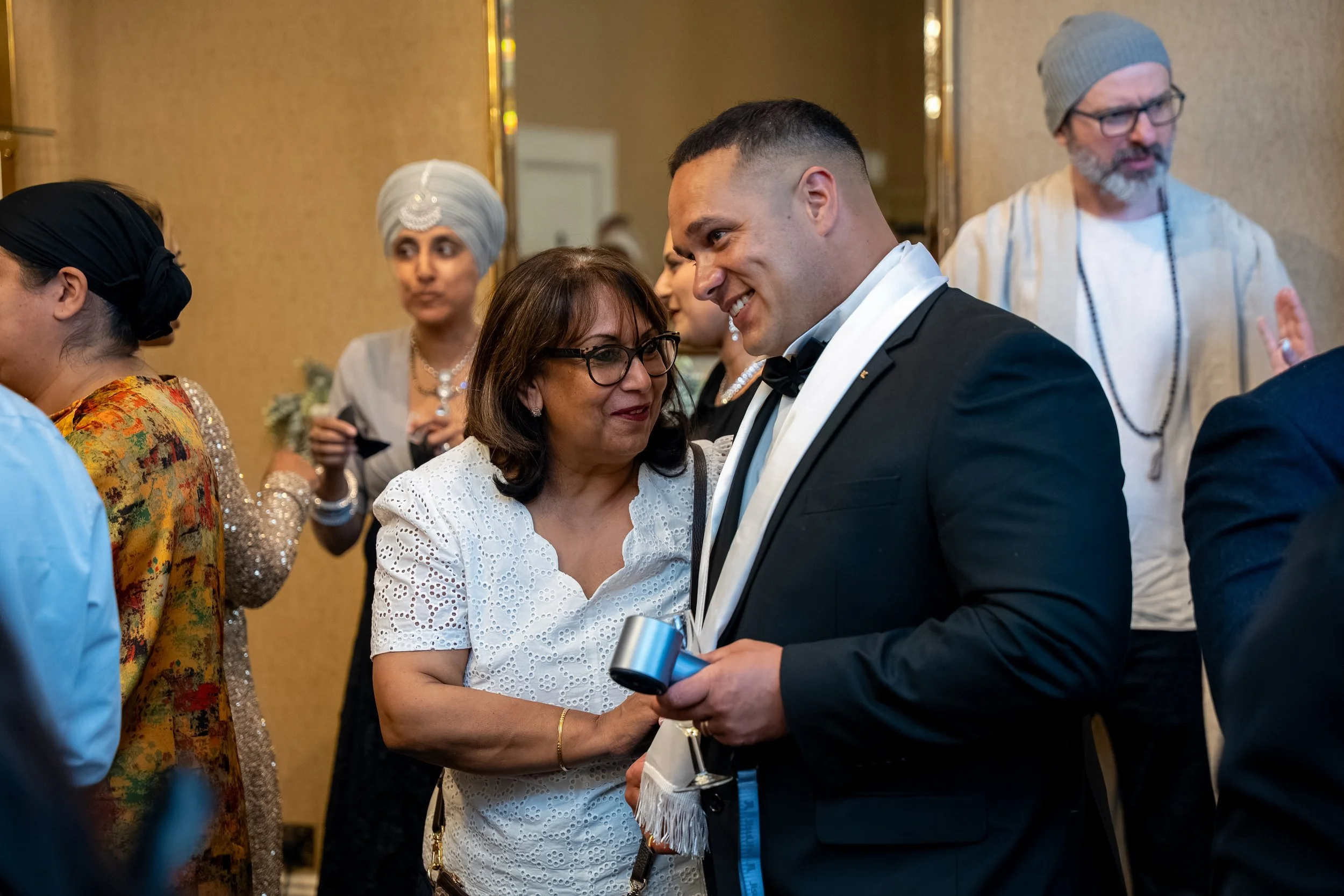 A group of diverse people at a formal event, exchanging greetings. A woman in white eyelet dress greets a man in a tuxedo, holding a glass of champagne, as other guests in traditional and formal attire look on.