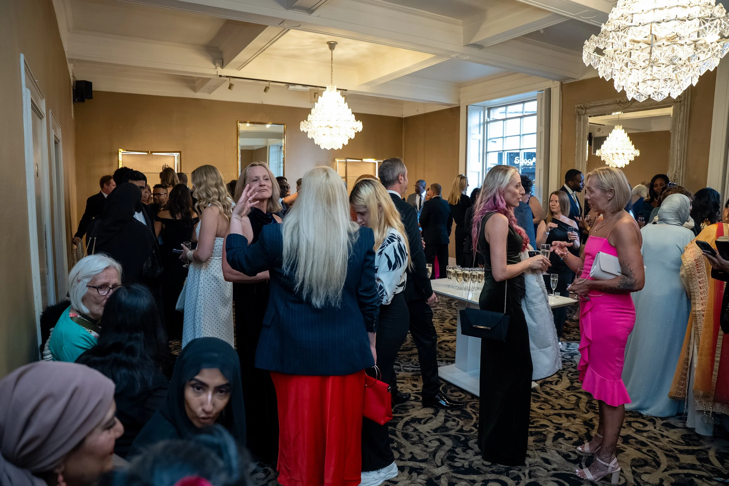 A crowded indoor event with people dressed in formal attire, mingling and conversing. The room has chandeliers, mirrors, and a patterned carpet.