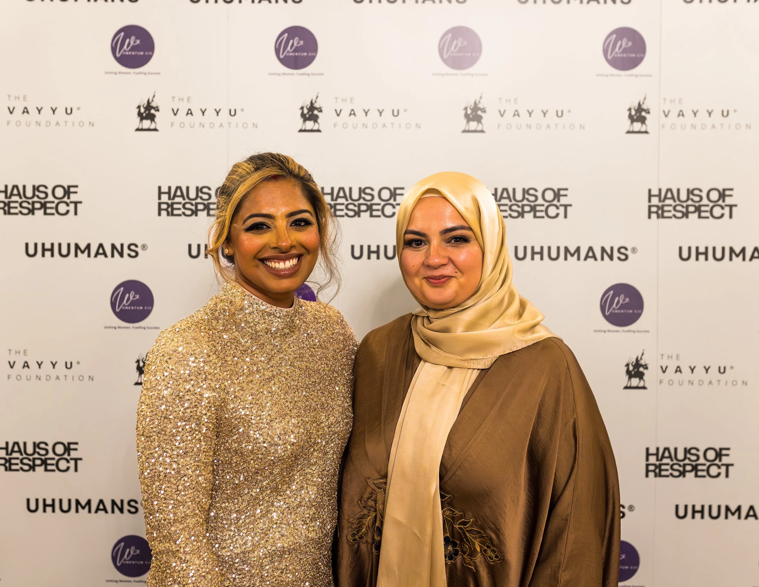 Two women smiling at a formal event, standing in front of a step-and-repeat banner with various logos and text, including 'The VAYYU Foundation', 'Haus of Respect', and 'UHUMANS'. The woman on the left has medium skin tone, blonde hair, and is wearin