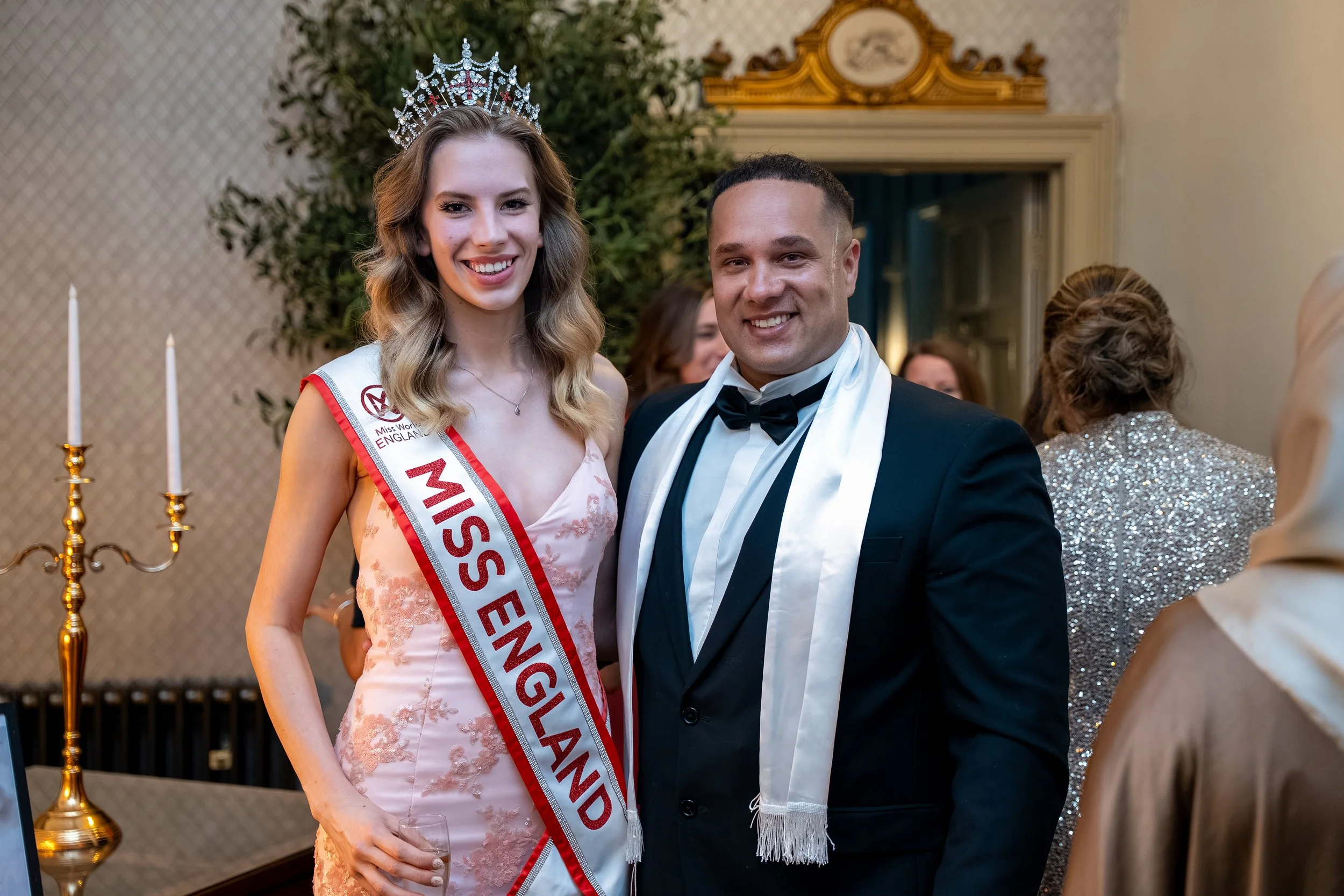 A woman wearing a Miss England sash and crown stands next to a man in a tuxedo at an indoor formal event. Both are smiling and looking at the camera.