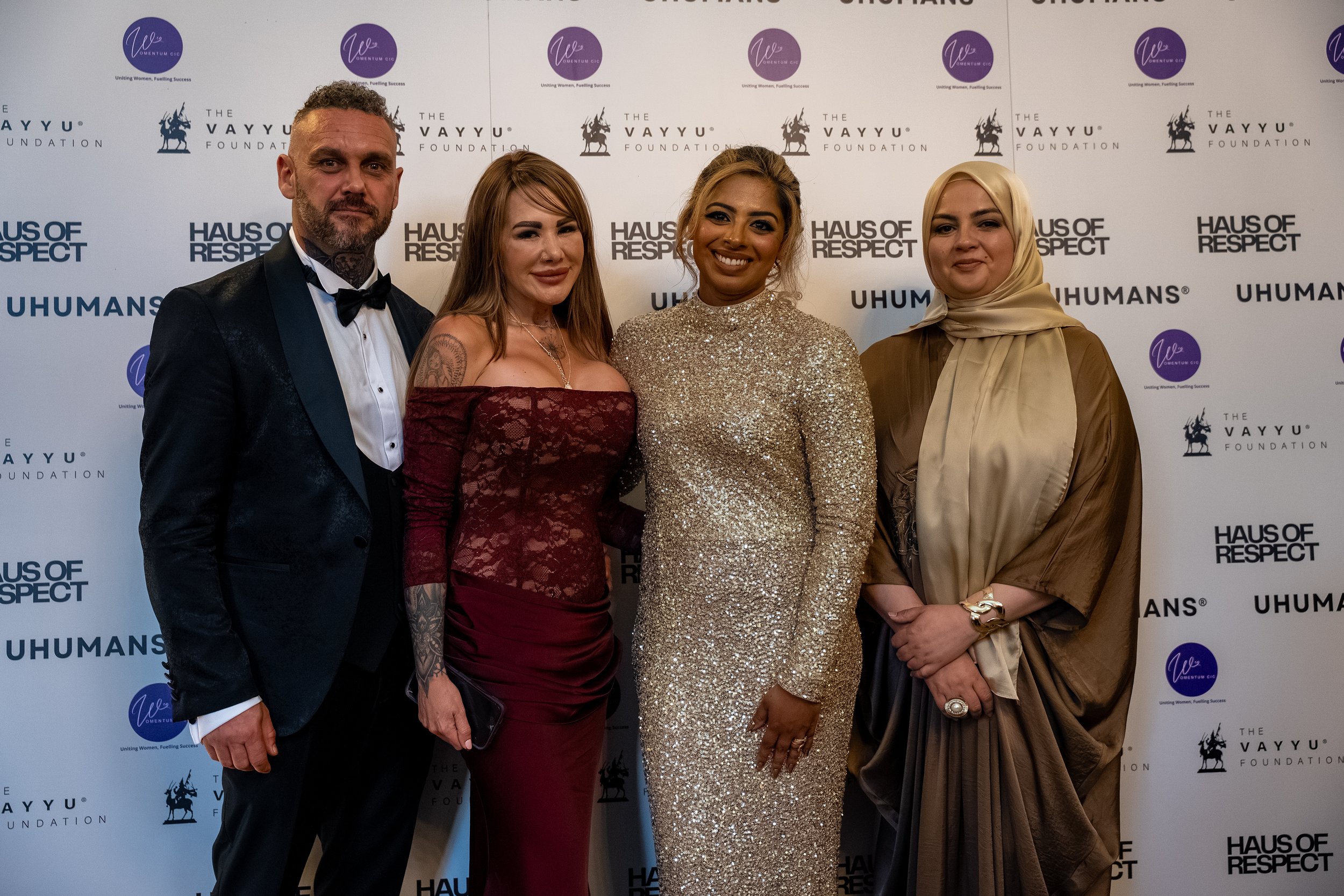 Four individuals dressed in formal attire standing in front of a white backdrop with logos related to VayyU Foundation, Haus of Respect, and Uhumans. The first person on the left is a man in a black tuxedo with a bow tie. The second person is a woman