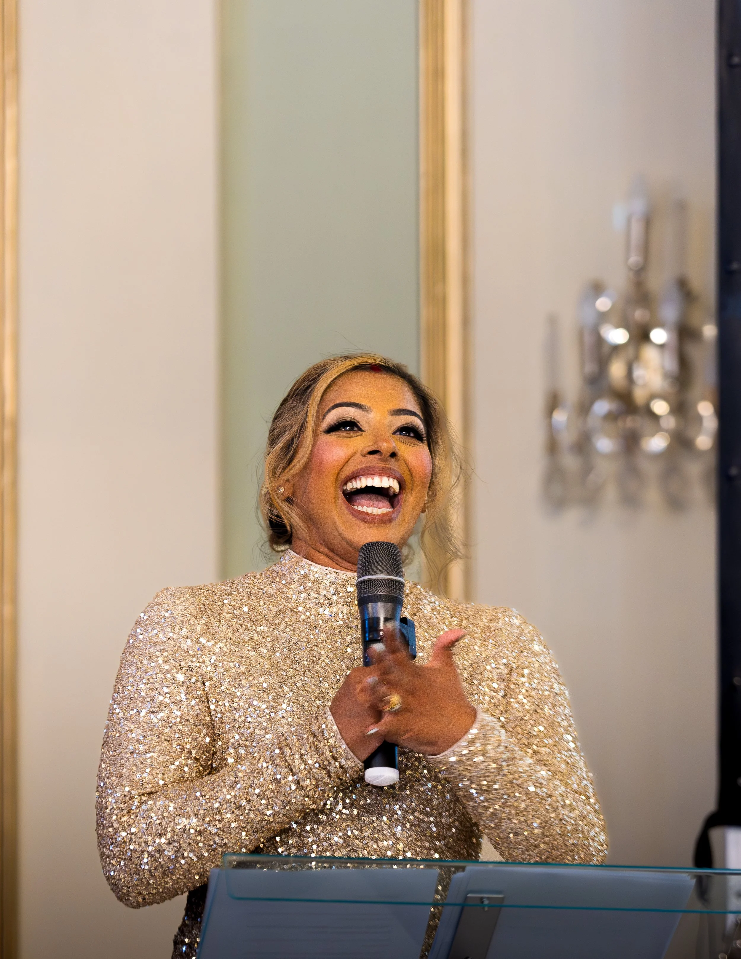 A woman smiling and laughing while holding a microphone, dressed in a gold sequined gown, standing in front of a music stand.