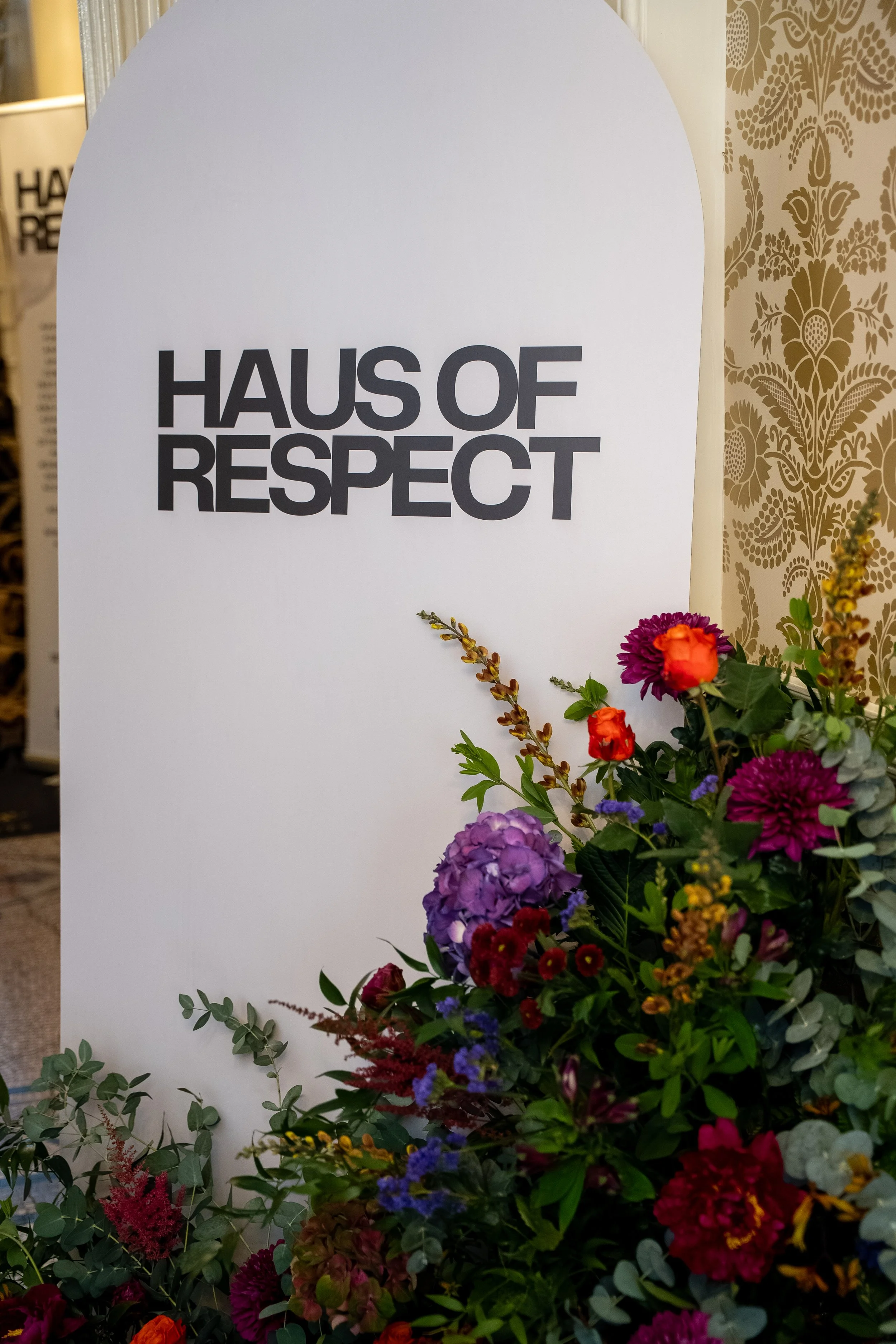 A white sign with the words 'Haus of Respect' in bold black letters, decorated with a flower arrangement of colorful flowers and green foliage in front of it.
