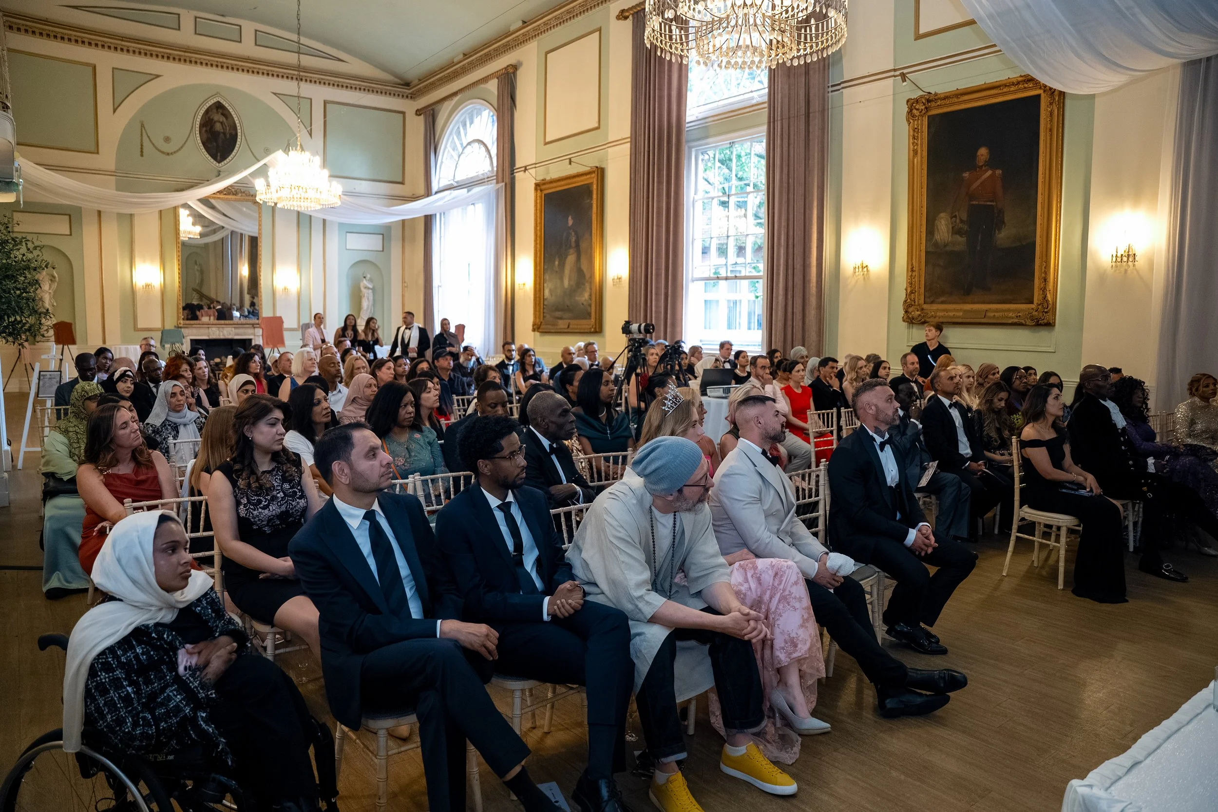 A large group of diverse people seated in a grand, elegant room with high ceilings, large windows, chandeliers, and gold-framed portraits, attending an event or presentation.