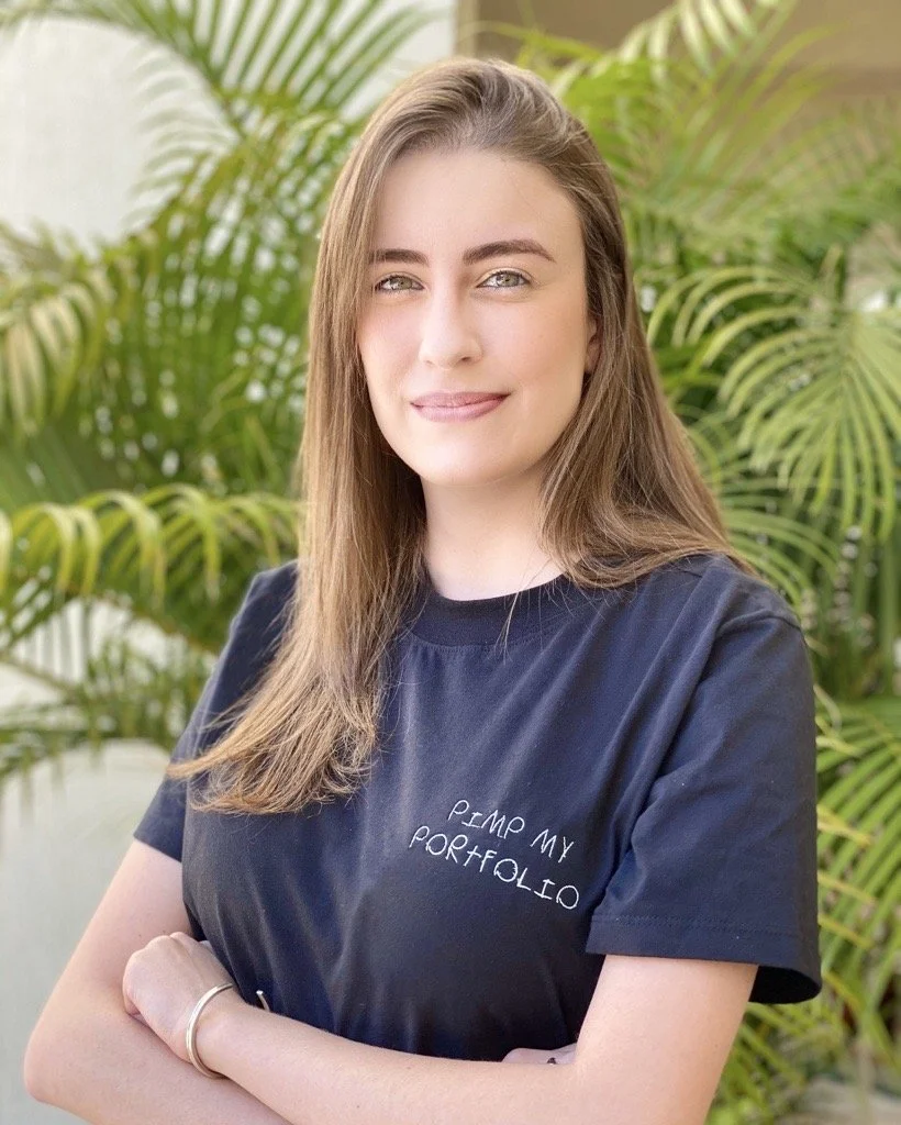 A young woman with long light brown hair and blue eyes stands outdoors in front of green tropical plants, smiling at the camera, wearing a black T-shirt that says 'PIMP MY PORTFOLIO' in white text.
