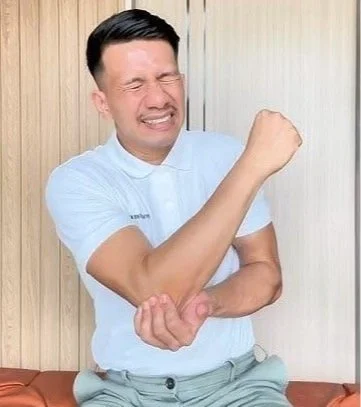 Hombre con expresión de dolor o incomodidad, cerrando los ojos y apretando el puño de un brazo mientras se apoya contra una pared de madera.