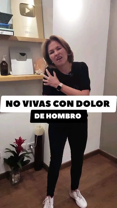 Mujer con expresión de dolor en el hombro, tocándose el hombro izquierdo, en un espacio interior con estantes y una planta