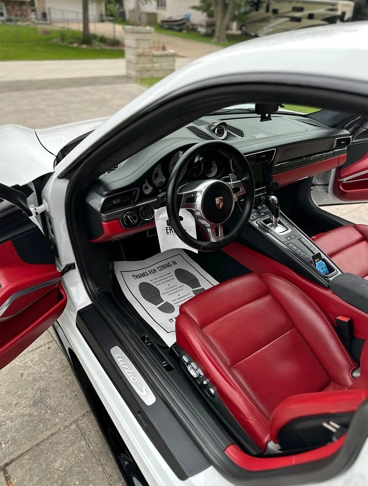 Inside a white Porsche 911 Turbo with red leather seats, displaying the dashboard, steering wheel, and gear shift, with a paper floor mat that reads 'Thanks for coming in'.