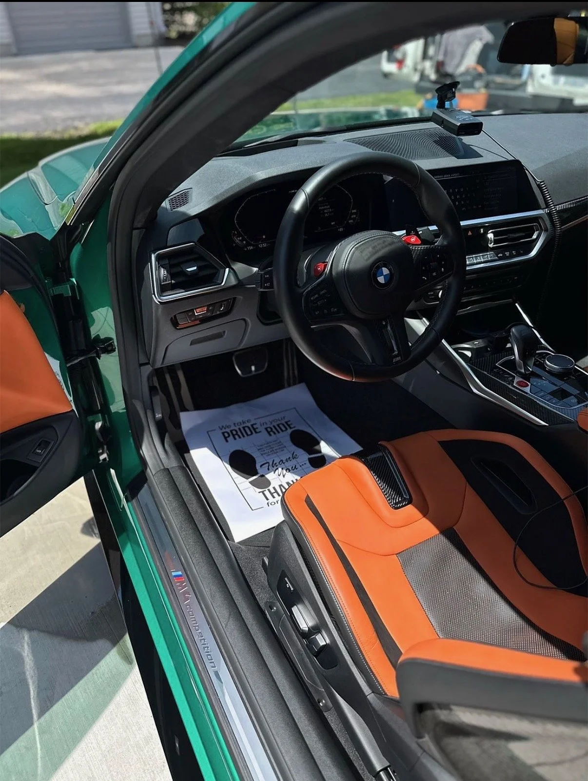 Interior of a BMW M vehicle with an orange and black seat, black steering wheel, digital dashboard, and a dashboard display. There is a note on the floor that mentions Pride Ride, thank you message, and footprints.