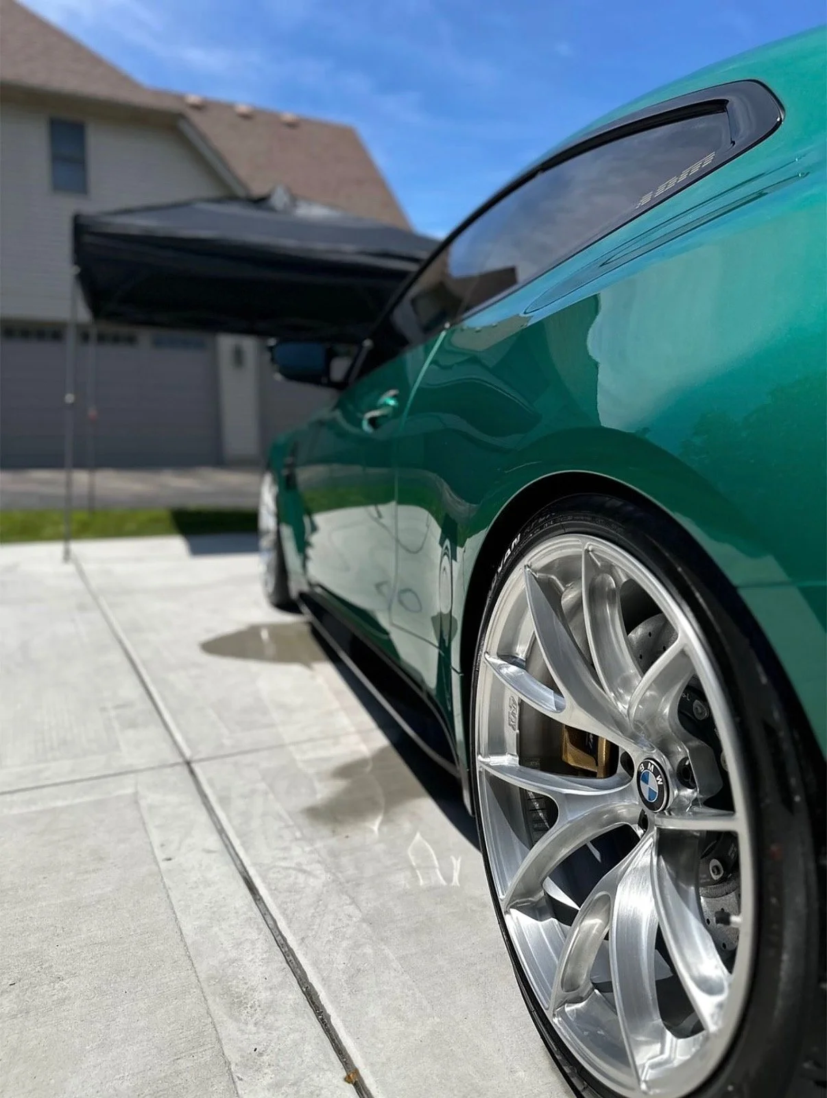 Close-up of a green BMW car with open hood, shiny alloy wheel, and residential driveway in the background under a blue sky.