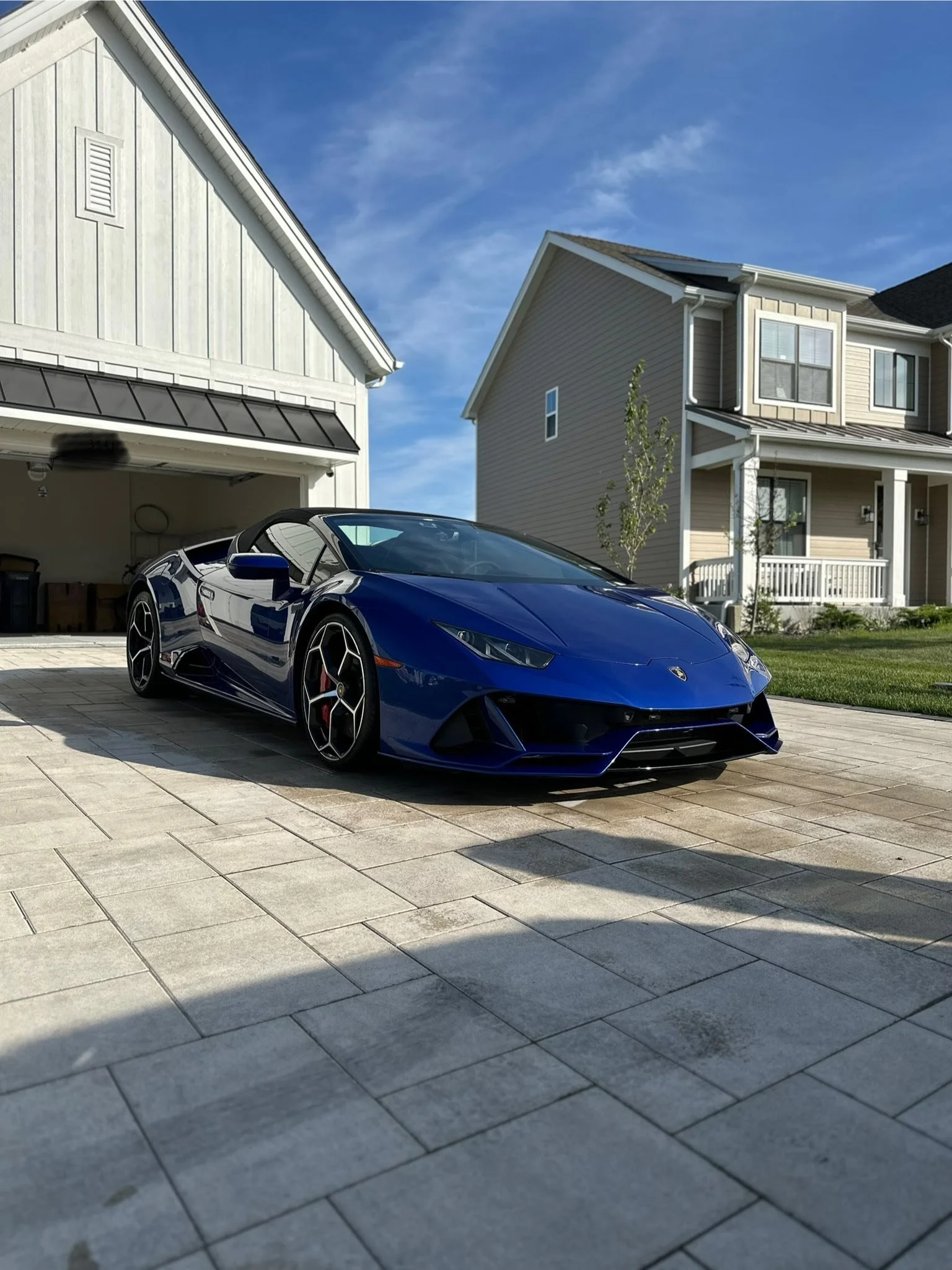 Blue Lamborghini sports car parked on a paved driveway in front of a house with a garage, neighboring house, lawn, and trees under a partly cloudy blue sky.