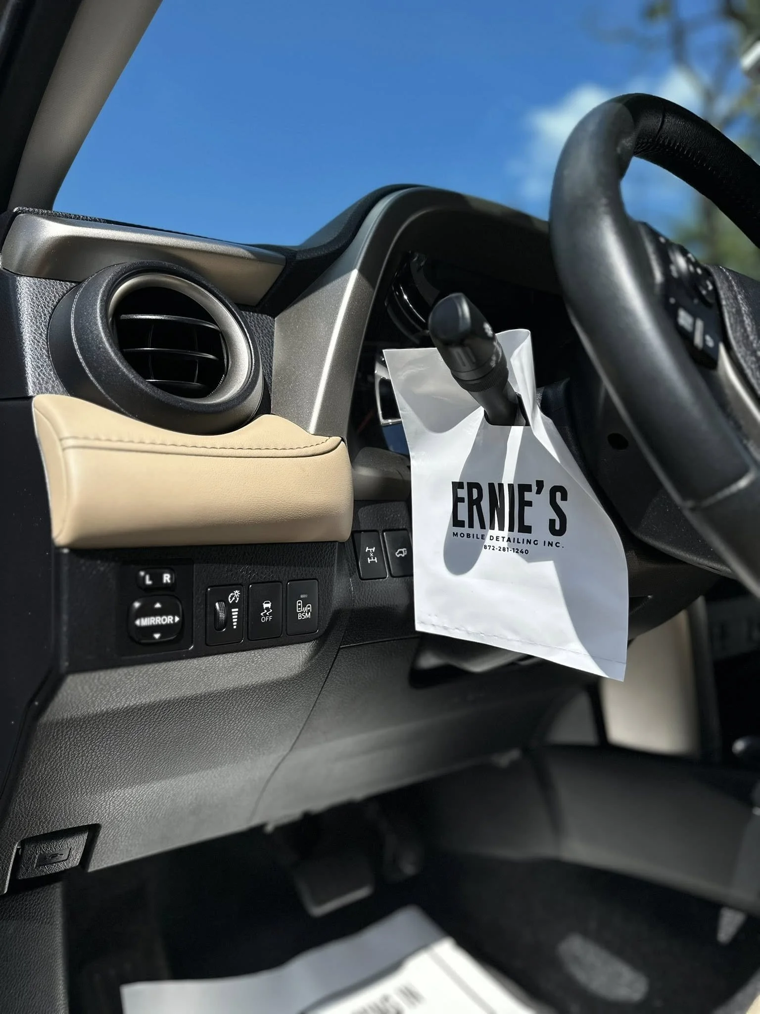 Close-up of a vehicle's dashboard showing an air vent, control switches, and a piece of paper with 'Ernie's' printed on it hanging from the steering column.