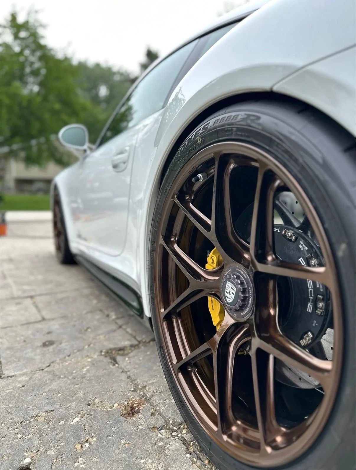 Close-up of a white luxury sports car with bronze alloy wheels, yellow brake calipers, and Porsche logo at the center of the wheel, parked on a stone-paved surface.