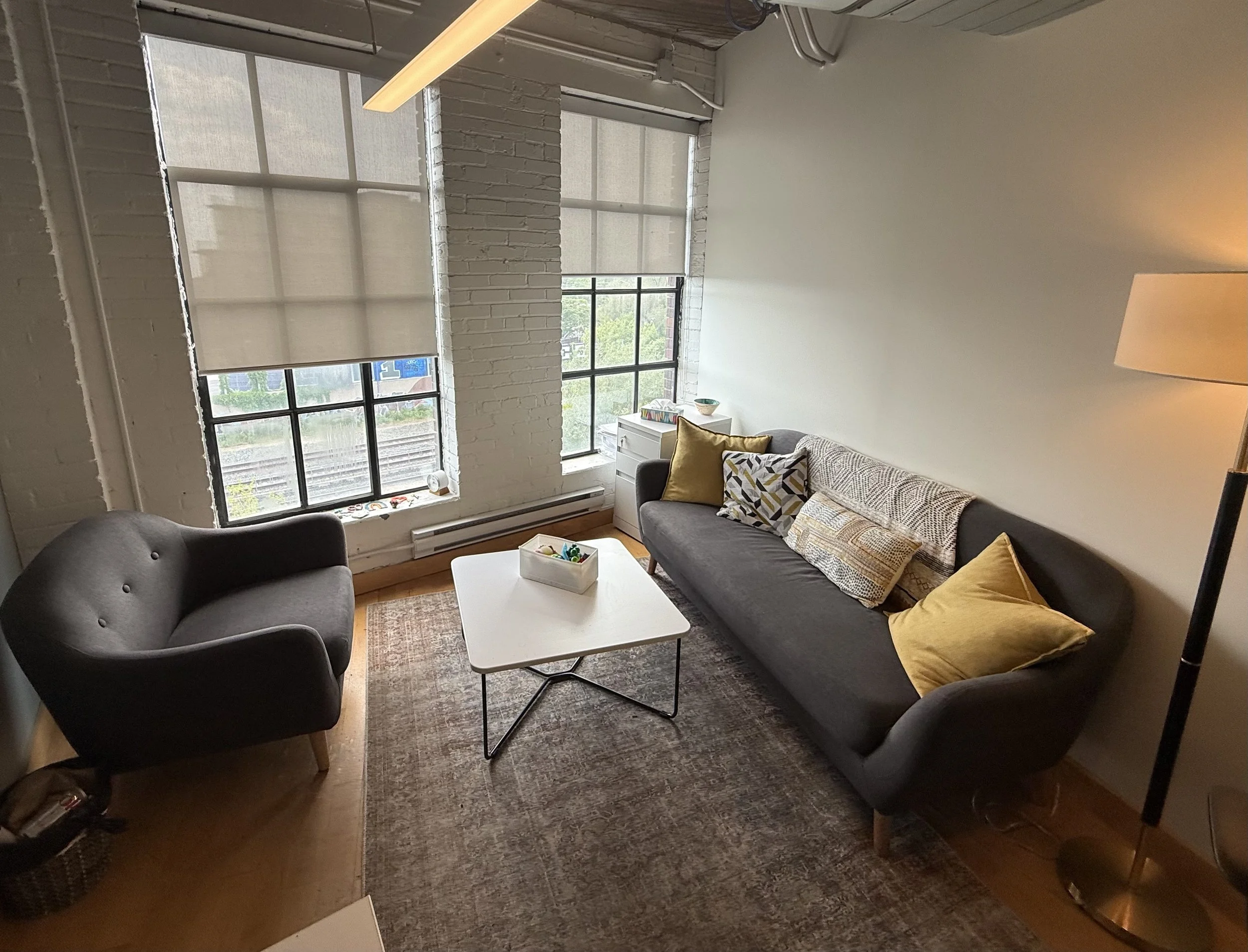 A modern living room with white brick walls, large windows with shades, a gray sofa with yellow and patterned pillows, a white coffee table with a box of colorful toys, a dark armchair, and a lamp in the corner.