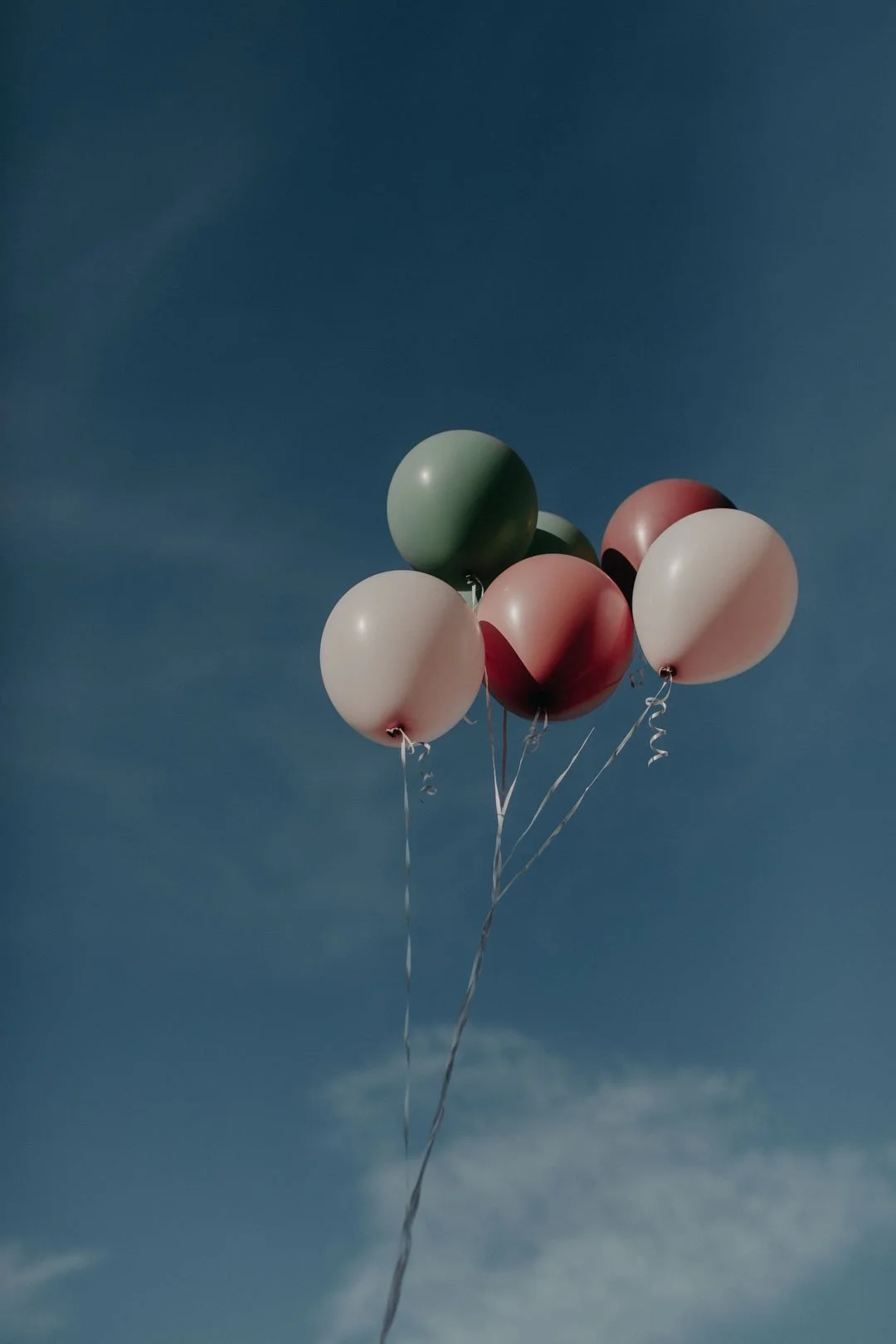 A bunch of six pastel-colored balloons, including green, pink, and white, floating against a blue sky with some wispy clouds.