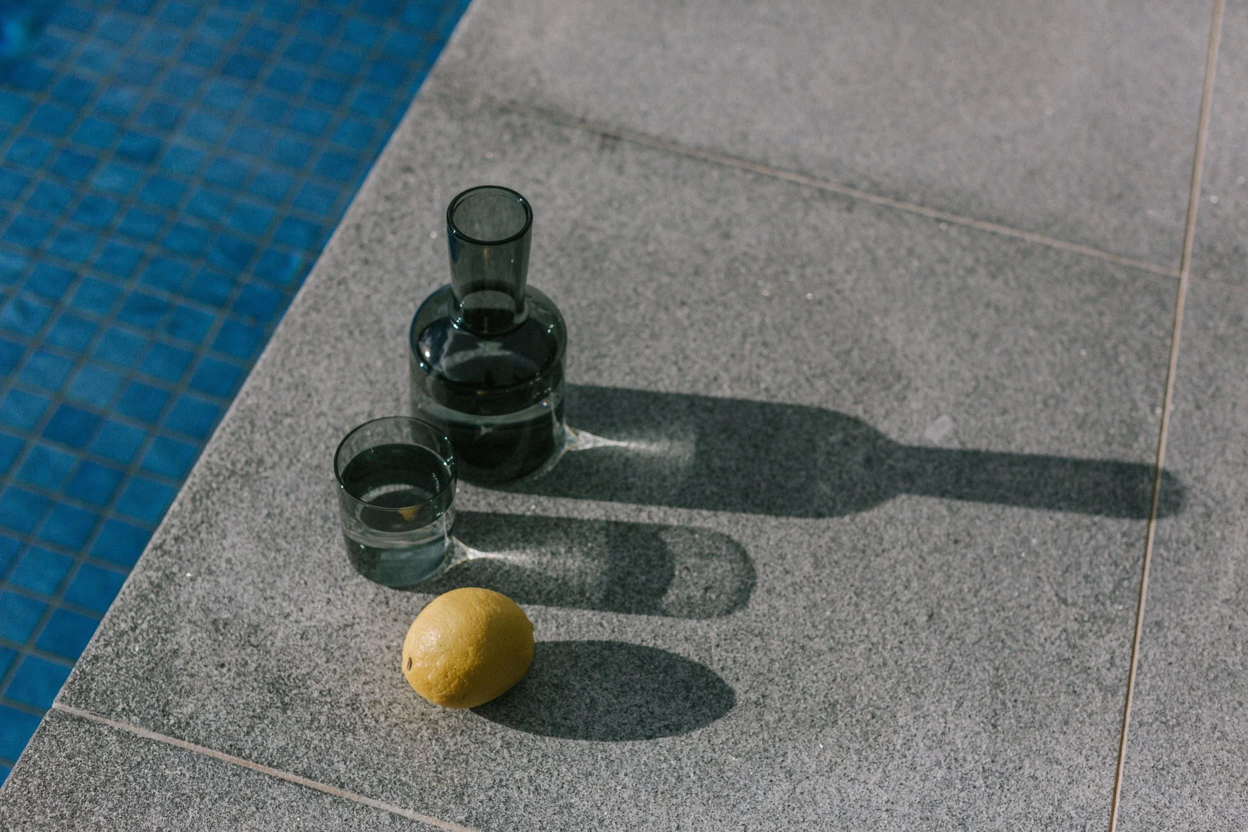 A green glass pitcher and a glass of water with a lemon on a concrete surface next to a swimming pool.