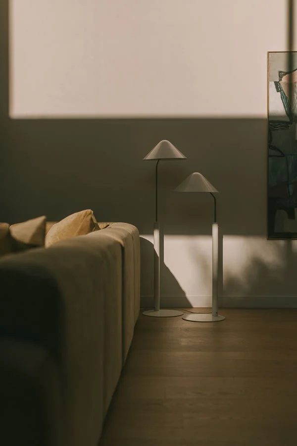 Living room with a beige sofa, two modern white floor lamps, and a large flat-screen TV mounted on the wall. Natural light creates shadows on the wall and floor.