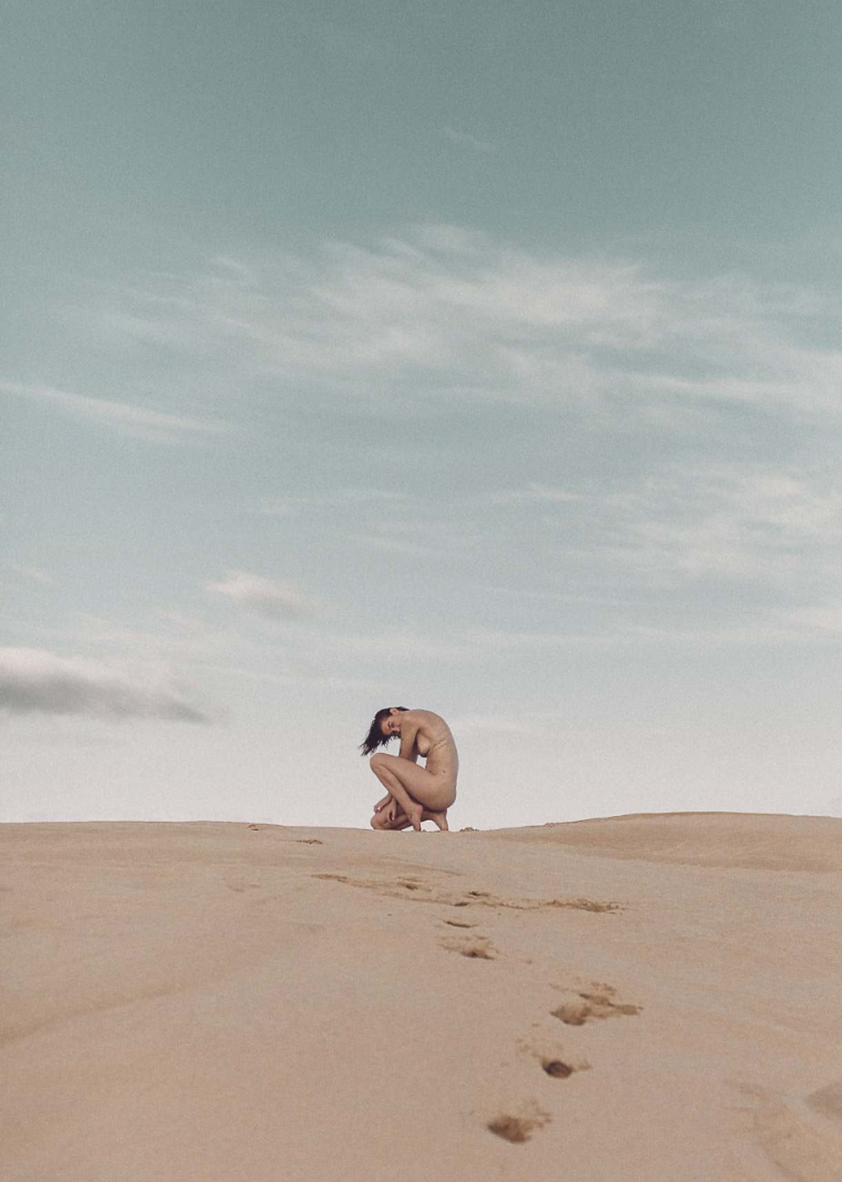 A person crouching on a sandy desert with footprints leading to them, under a cloudy sky.
