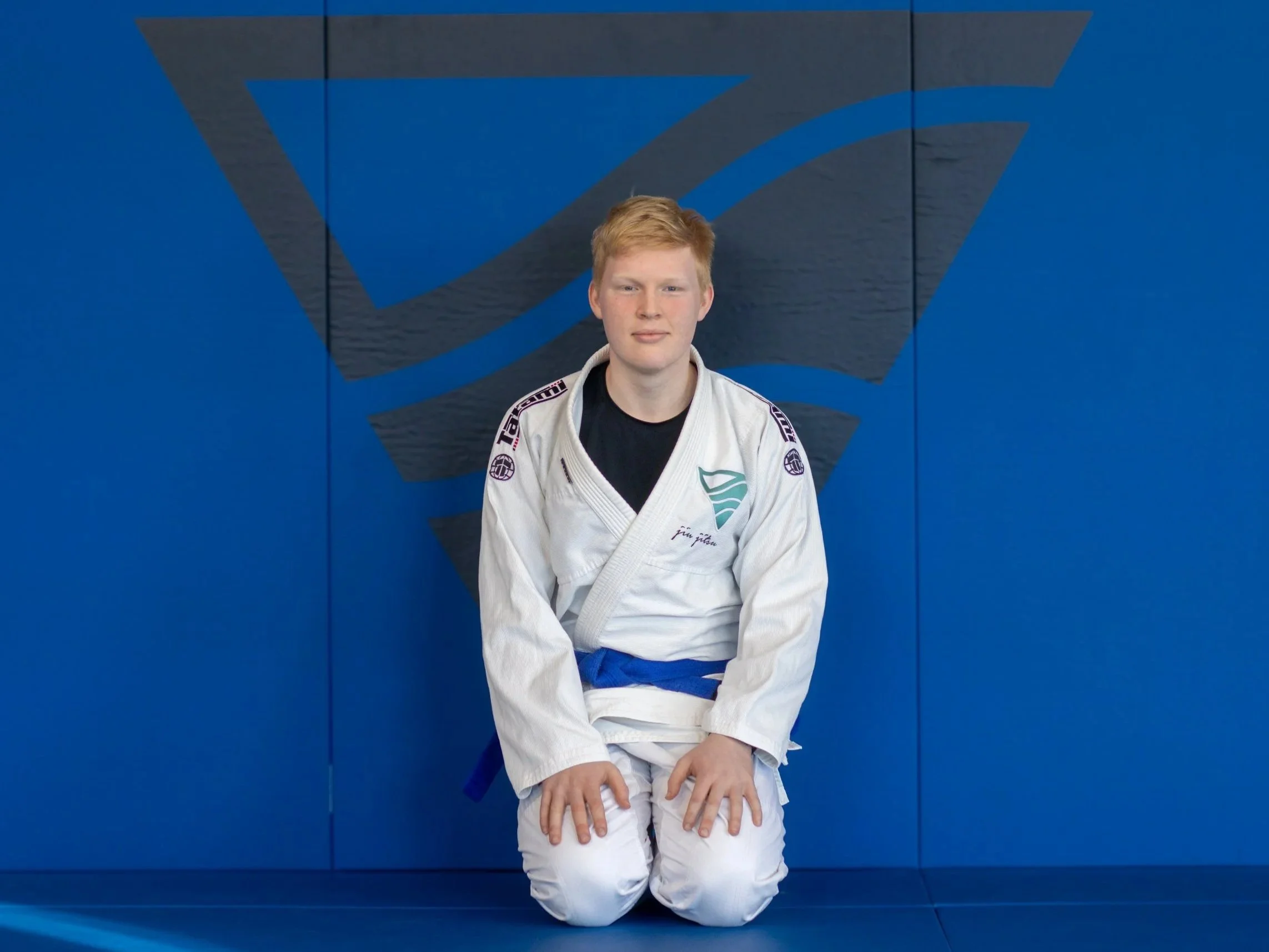 A young man with red hair wearing a white Brazilian Jiu-Jitsu gi and blue belt, sitting against a blue wall with a black and blue abstract graphic design.