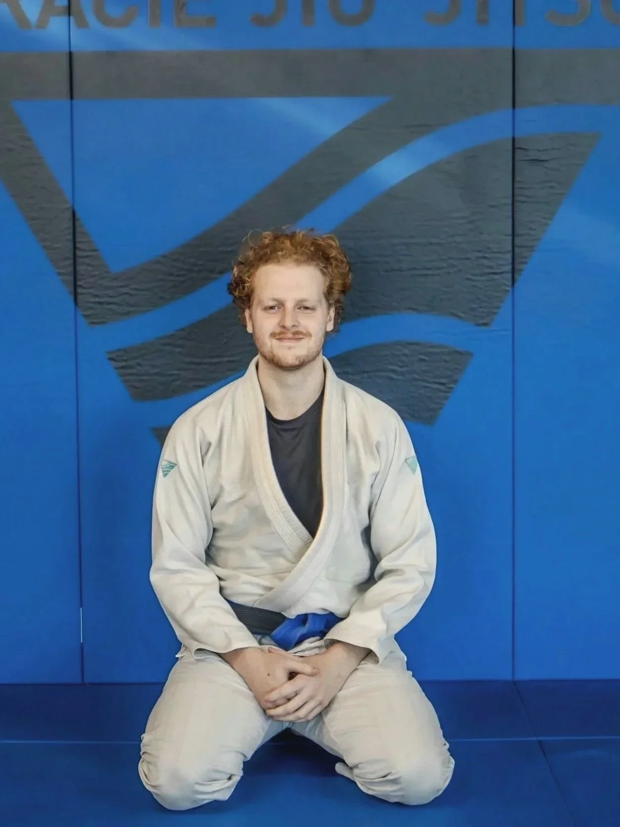 A man with curly red hair and a beard sitting in a kneeling position in a Brazilian Jiu-Jitsu gi in front of a blue wall with a large black and blue logo and partially visible text.