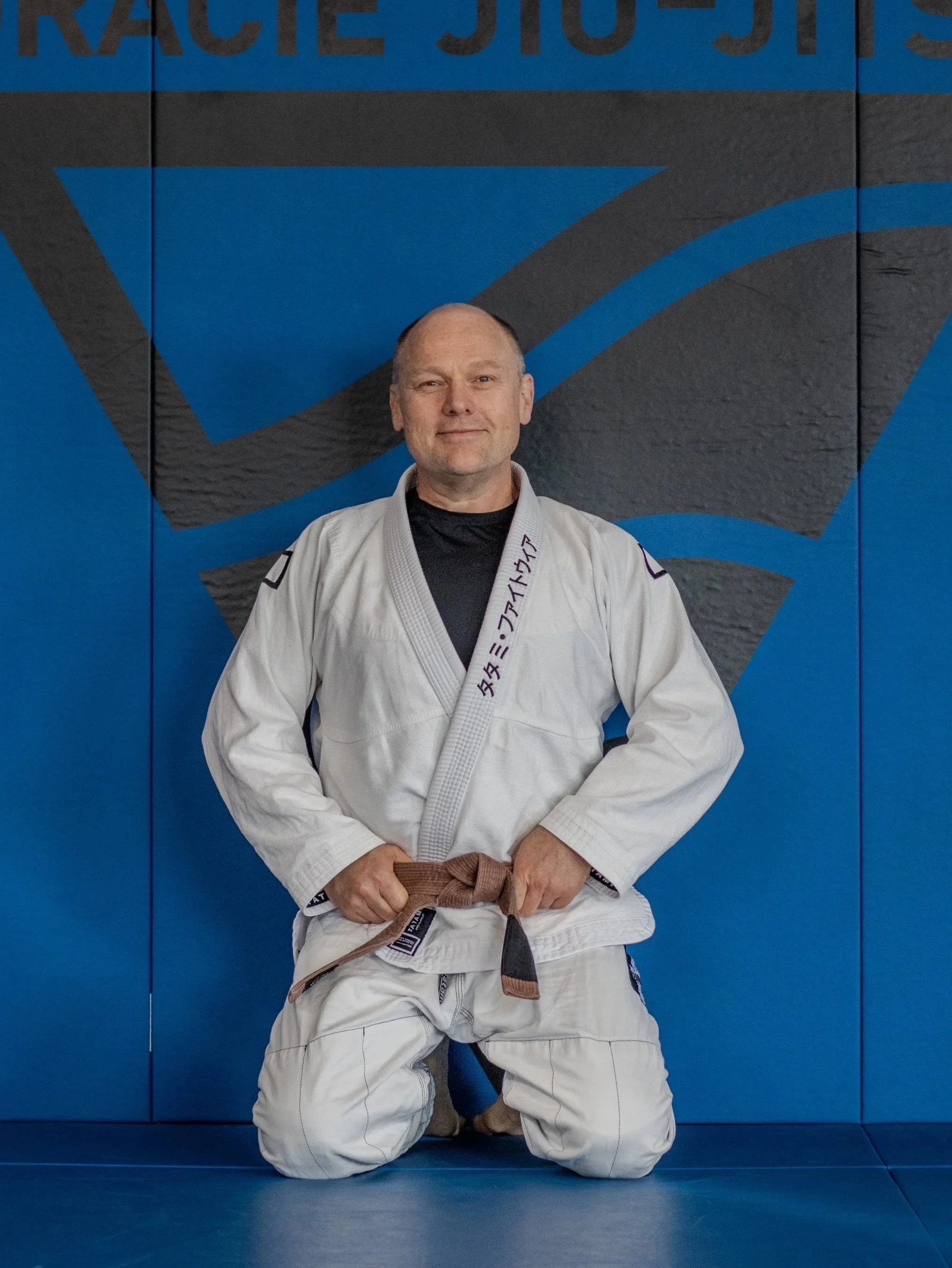 A man in a white Brazilian Jiu-Jitsu gi with a brown belt, kneeling on a blue mat in a gym, with a large blue and black mural behind him.