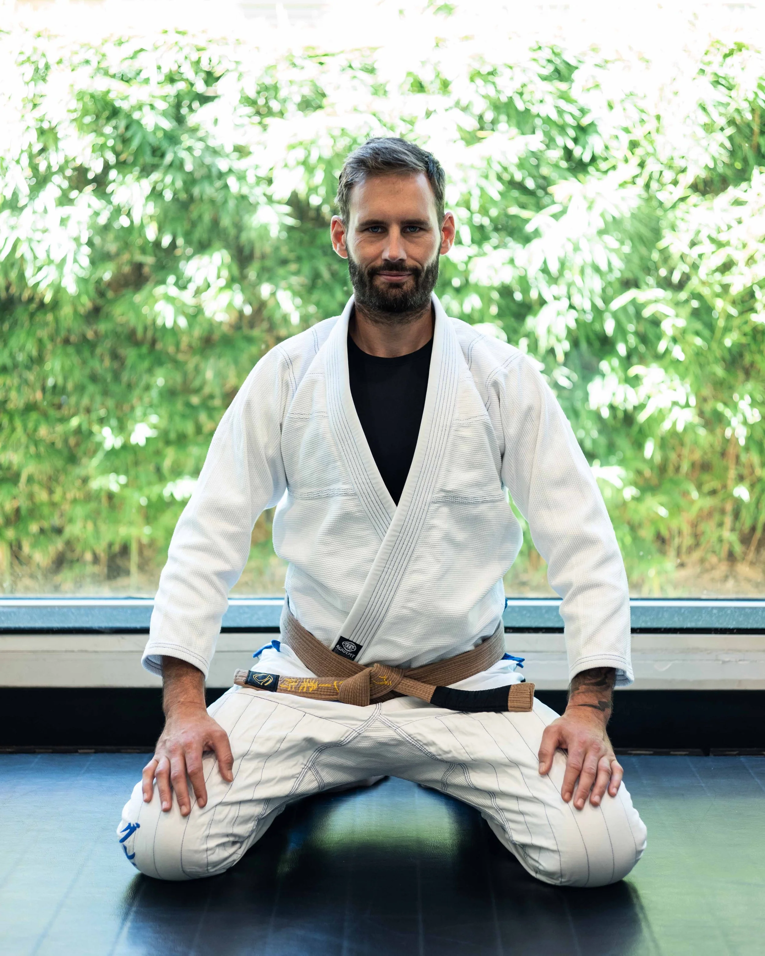 A man with a beard and short dark hair sitting on a martial arts mat in a gi, with a brown belt, in front of a window and greenery outside.