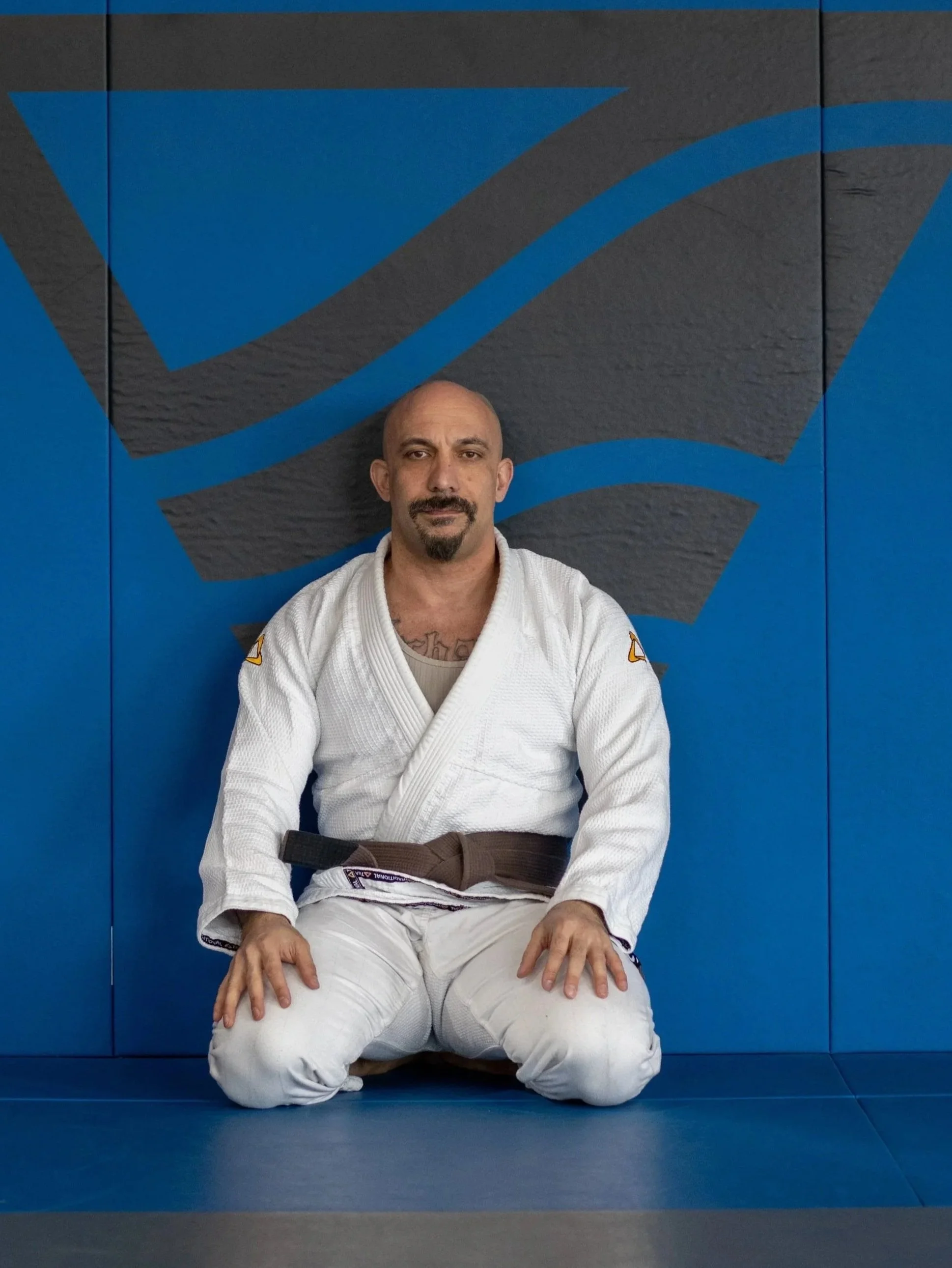 A man in a white Brazilian Jiu-Jitsu gi sitting on his knees in a dojo with blue mat walls behind him.