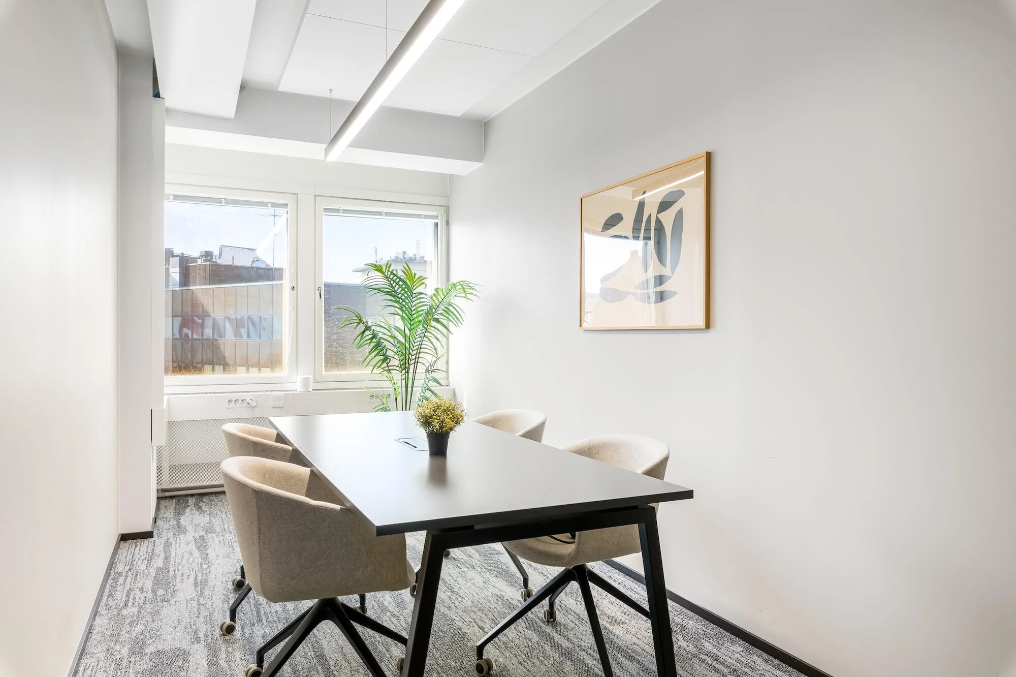 Boffice Sörnäisten kokoushuone, jossa valkoinen pöytä, viisi teollisuustyylistä tuolia, kiiltävä televisioruutu, taulut seinällä ja suuri kasvi lattialla