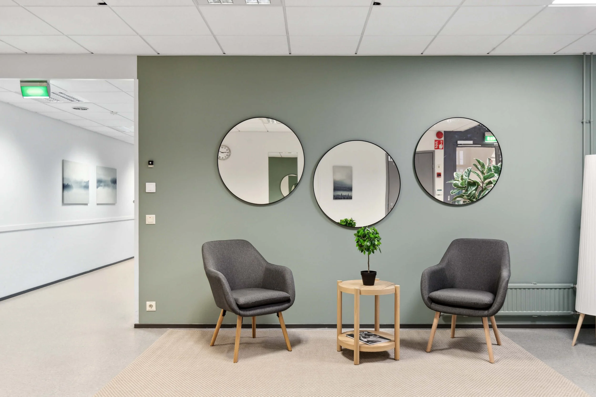 Reception area with two grey chairs, small table with small plant and leaves, three round mirrors and two murals on the back wall, partial door and window.