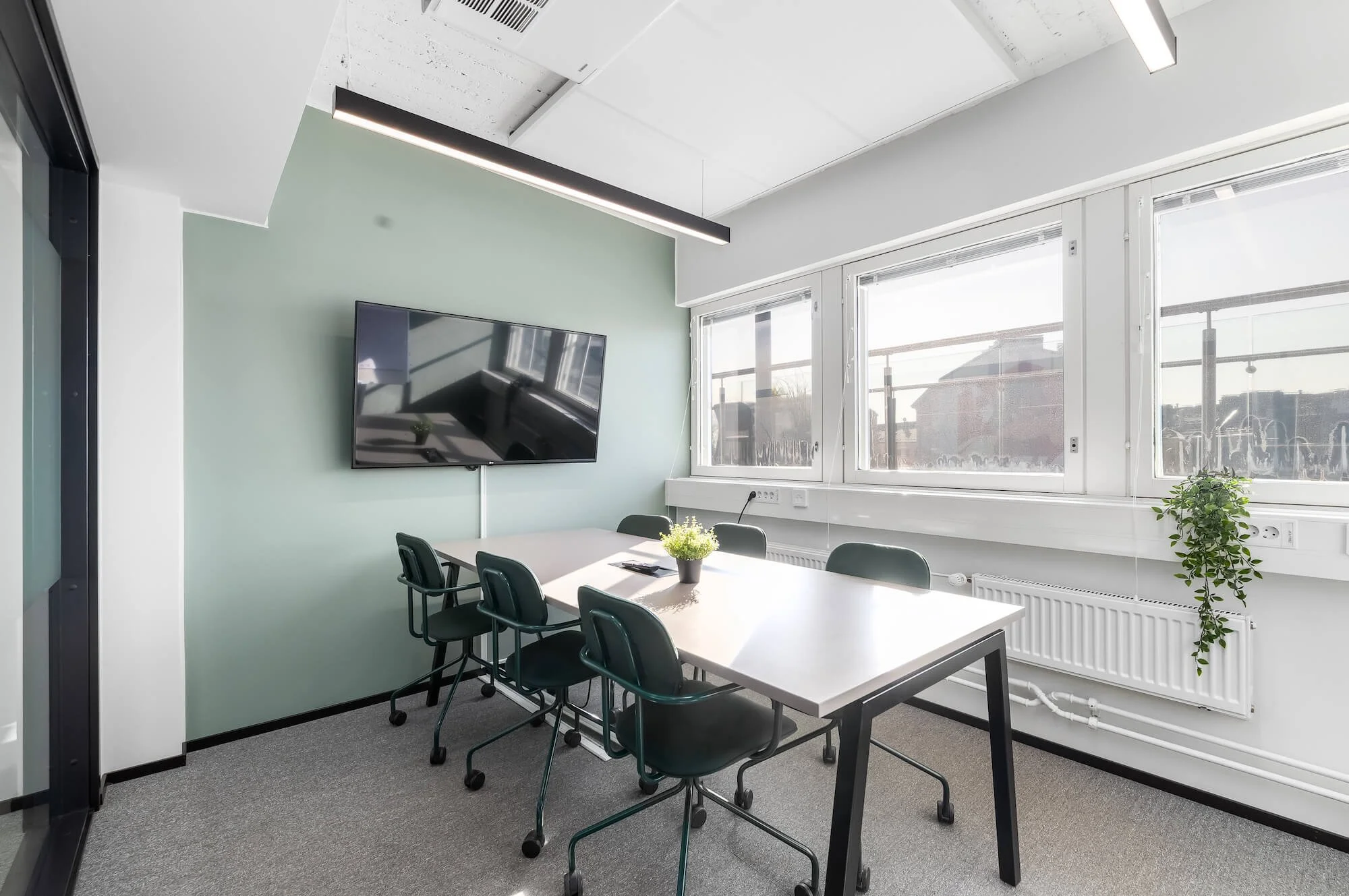 A photo of the interior of the Boffice Sörnäinen meeting room, featuring a round table, six dark chairs, a large TV on the wall, and large windows offering a view of trees and part of the building.