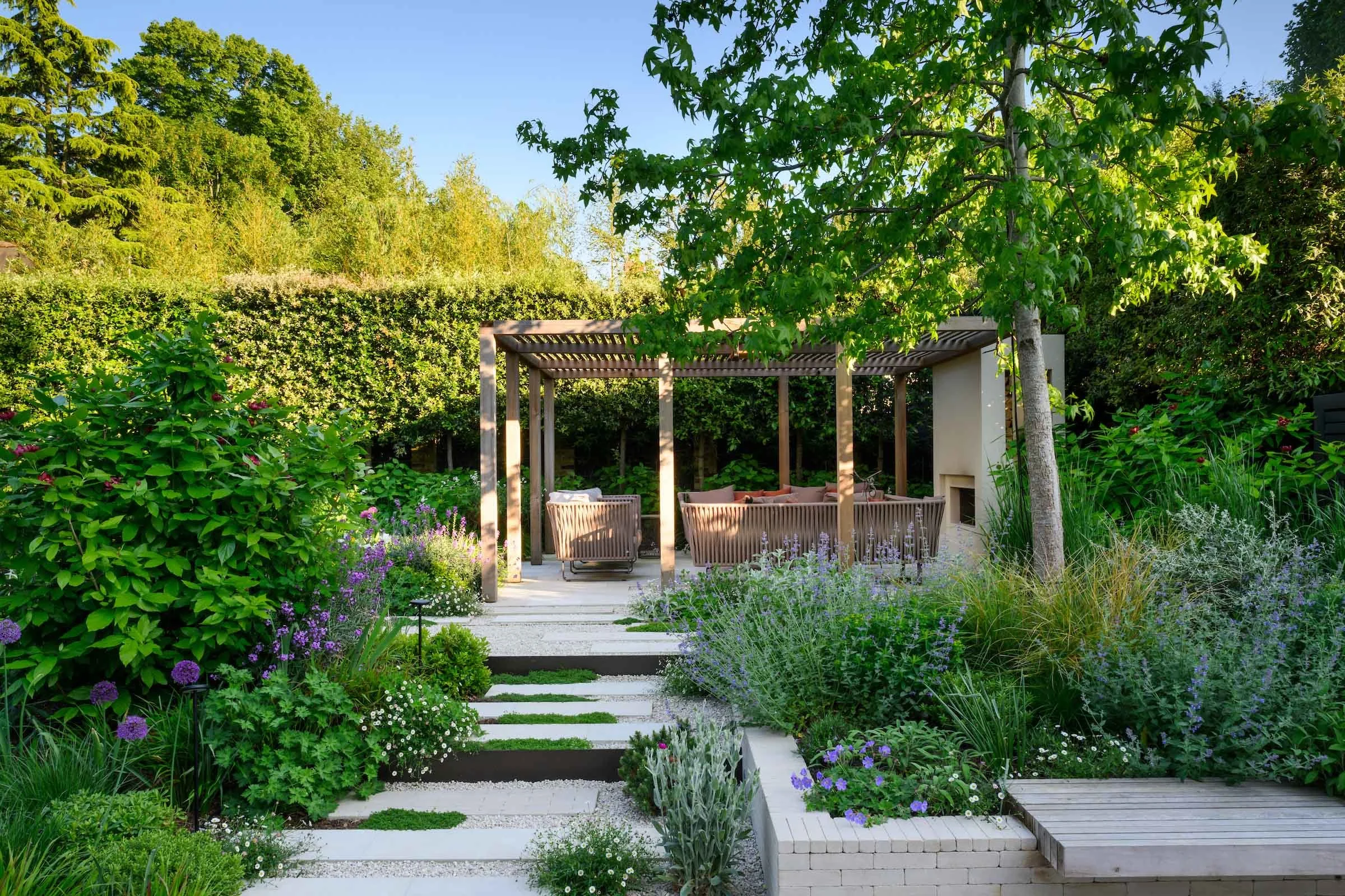 Timber pergola with seating, limestone steps and thyme-planted rills among lush borders in a Putney garden by Kawauchi Rowe